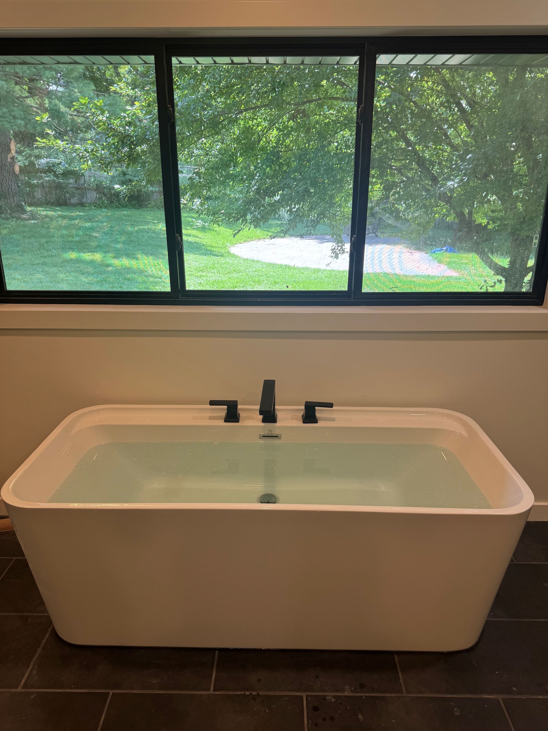 Modern freestanding bathtub in a tiled bathroom beneath a large window overlooking greenery