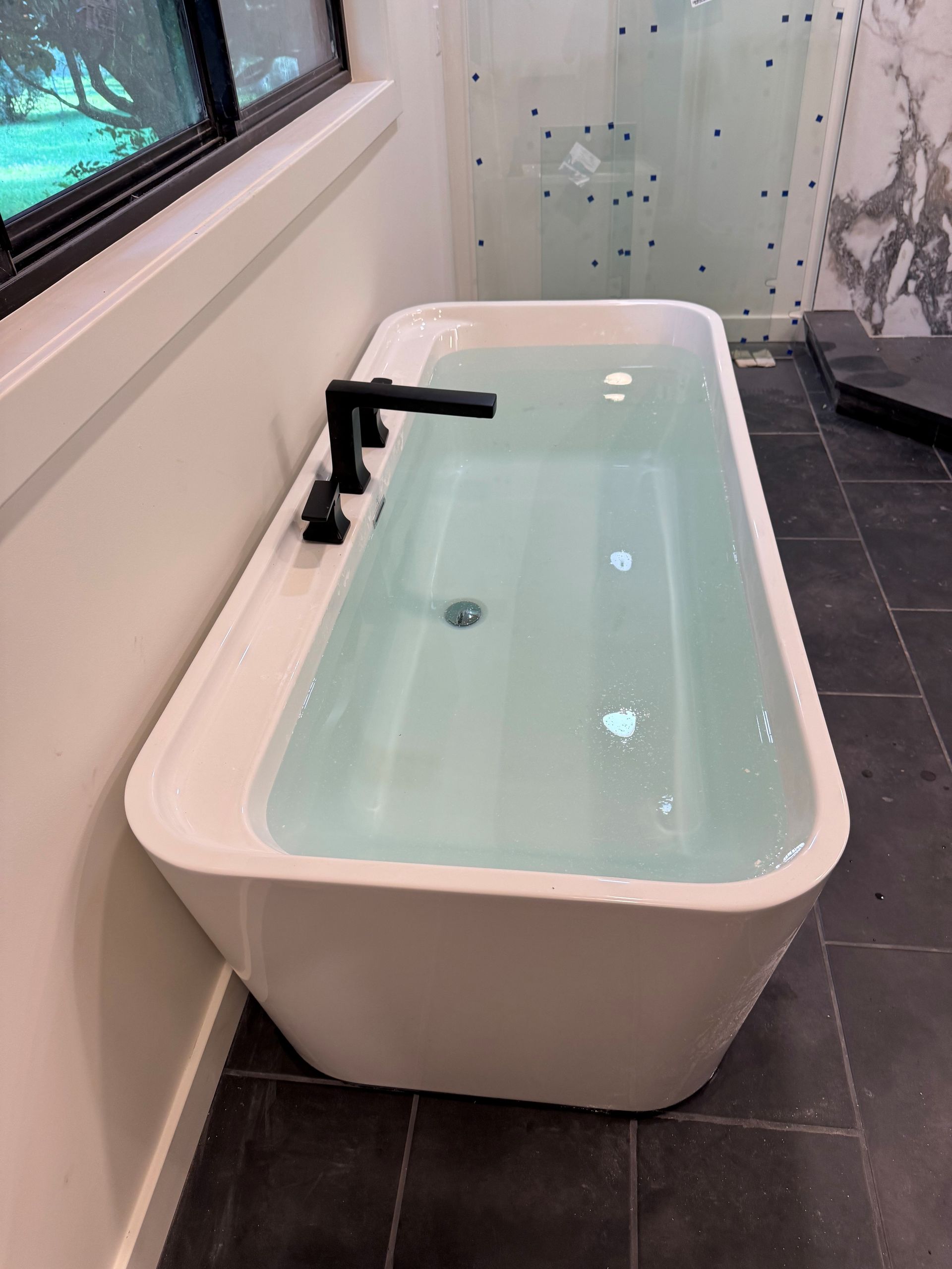 Modern white bathtub with black faucet in a tiled bathroom, filled with pale green water.