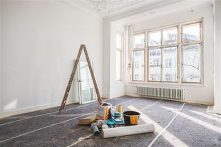 Bright empty room with a ladder, paint supplies, and rolled carpet by a large window