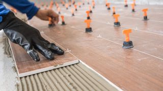 Gloved hand spreading adhesive on floor tiles during installation of orange leveling clips