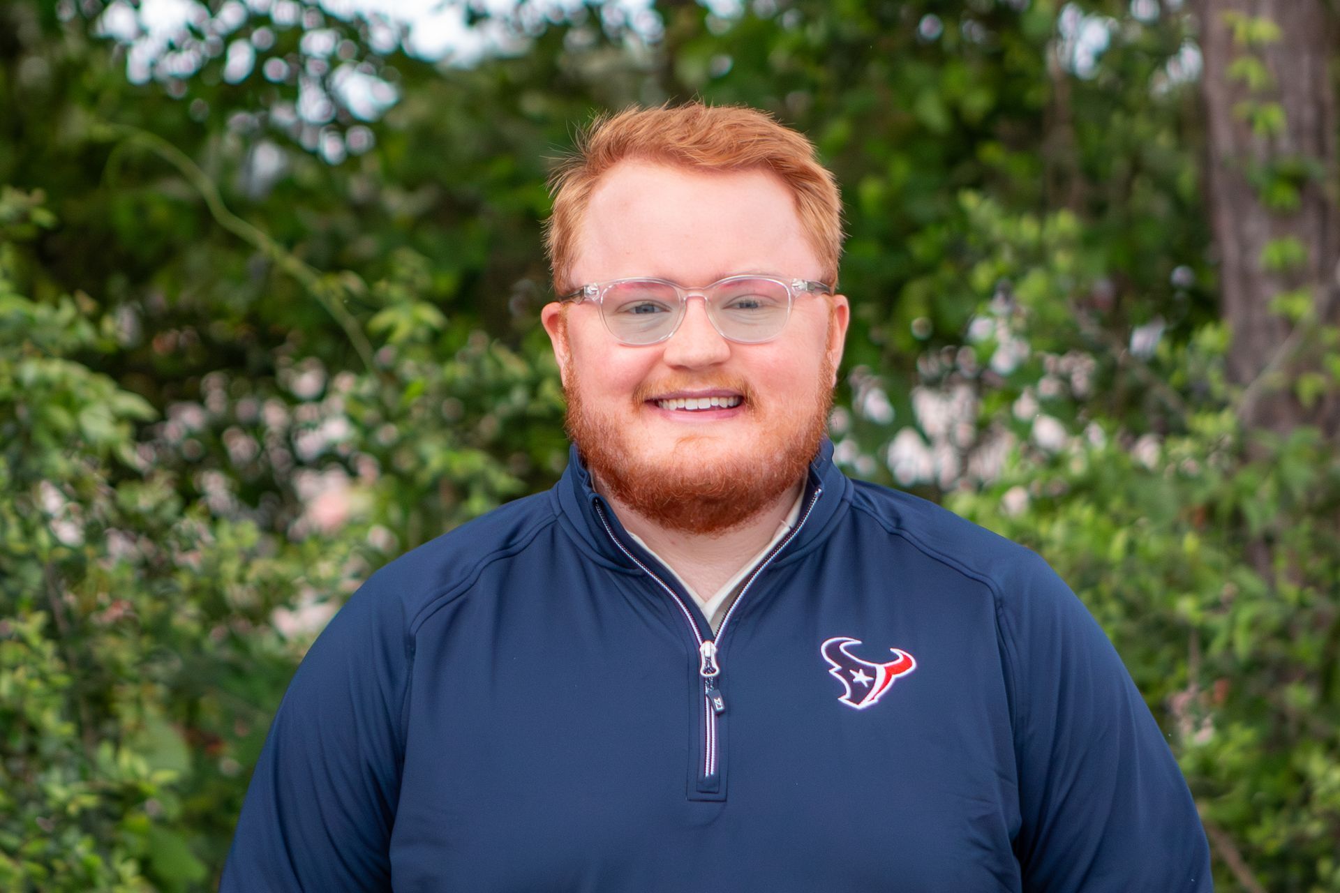 Smiling man in a navy pullover standing outdoors with green trees in the background