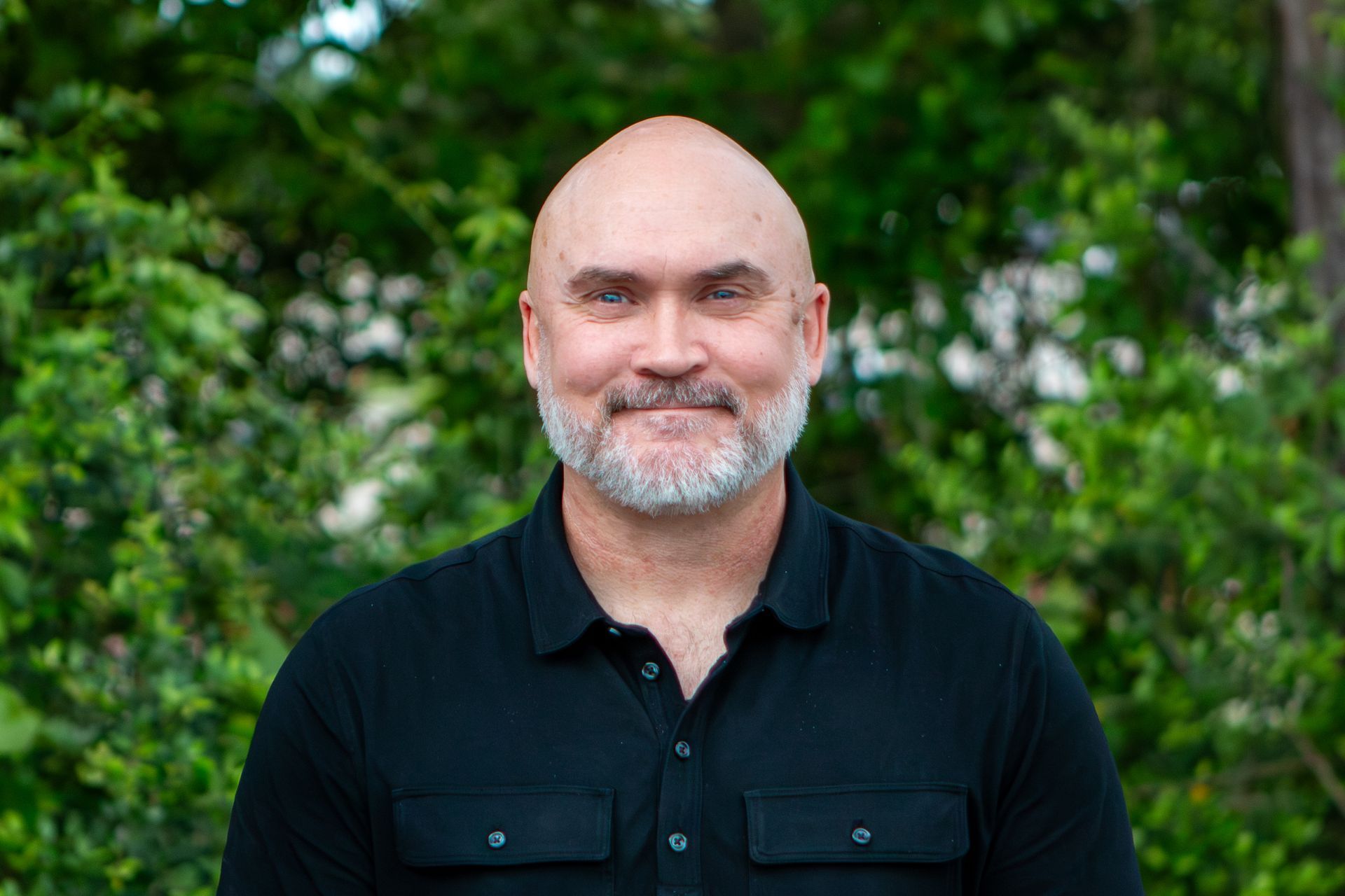Smiling bald man in a black shirt standing outdoors with green foliage behind him