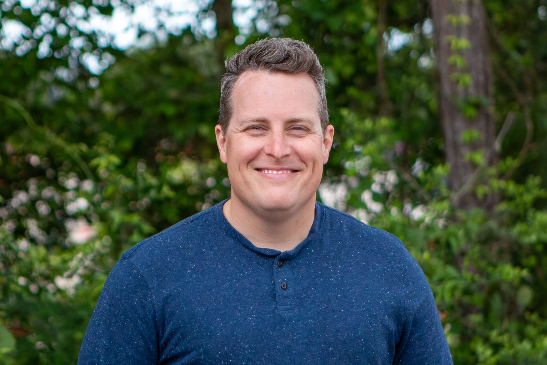 Smiling man in a blue sweater standing outdoors with trees in the background