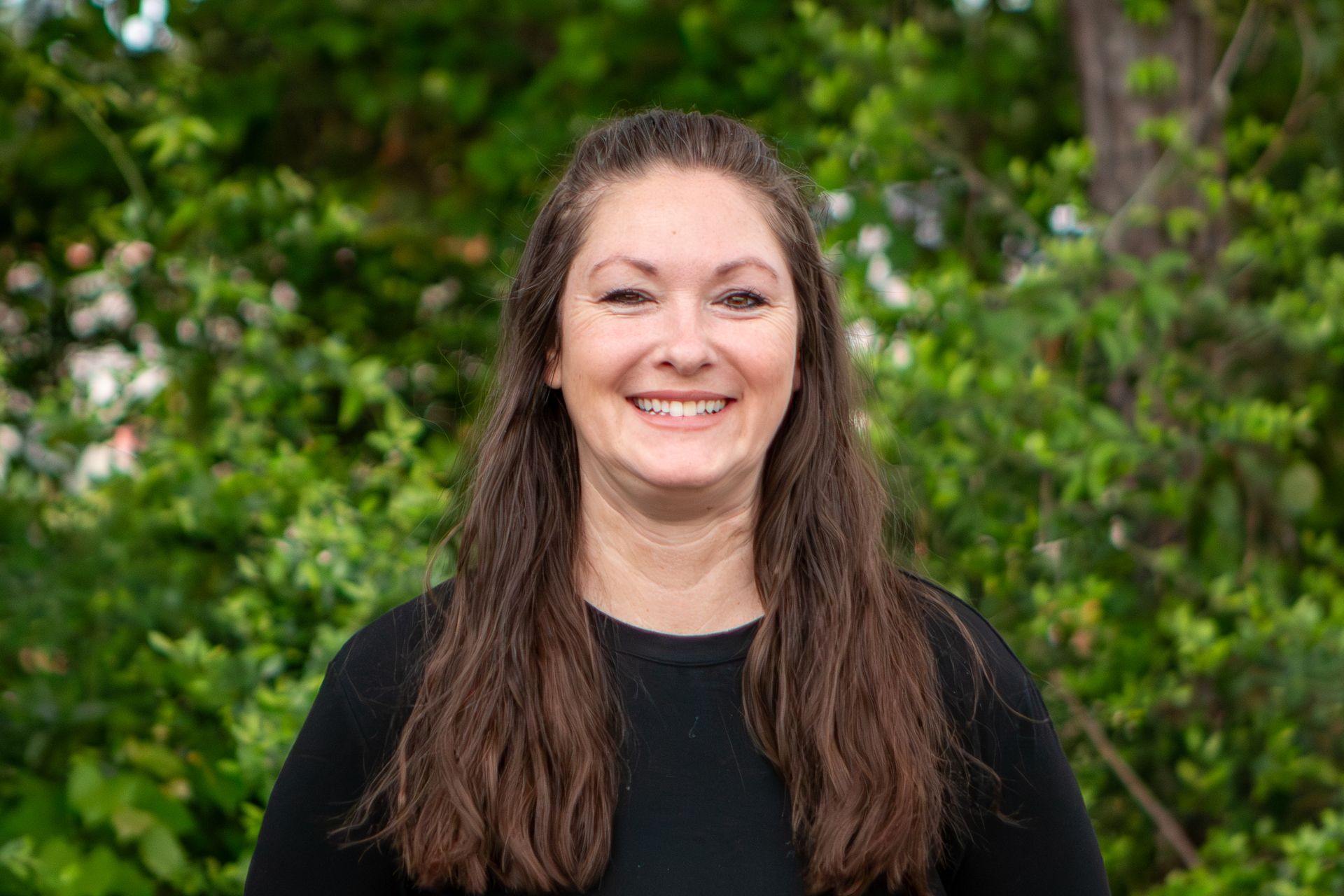 Smiling woman with long brown hair standing outdoors in front of leafy green trees