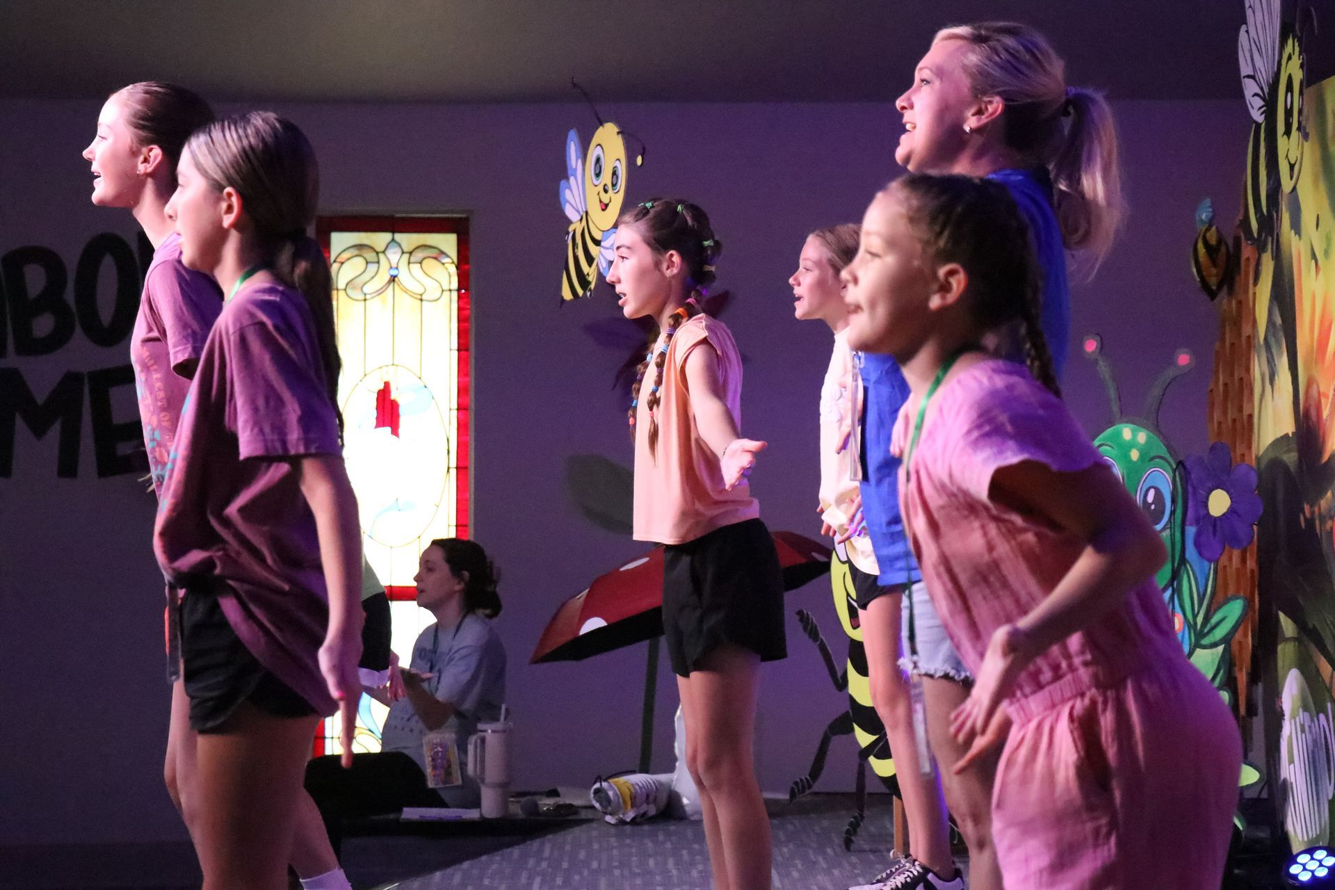 Children rehearsing on a stage with purple lighting, arms raised in a dance pose