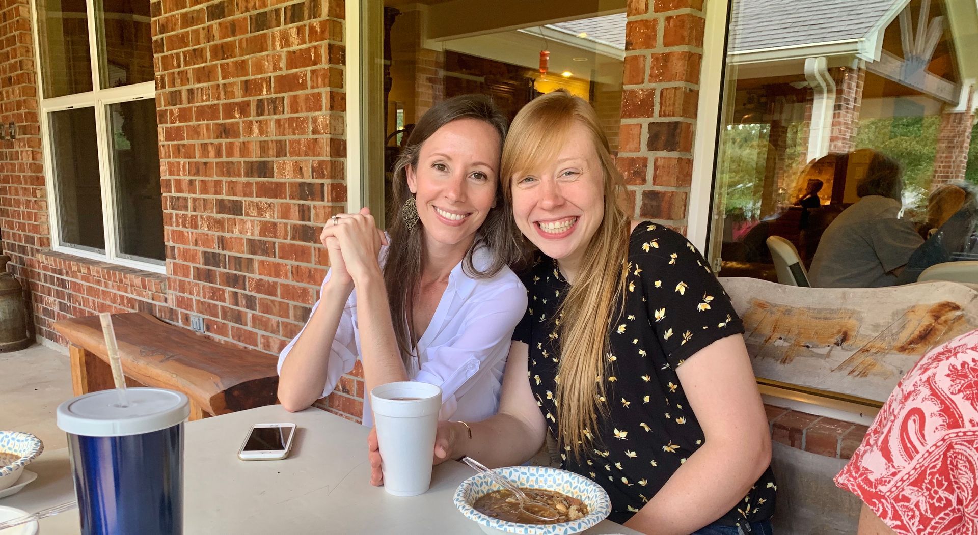 Two smiling people seated at an outdoor café table with drinks, in front of a brick wall