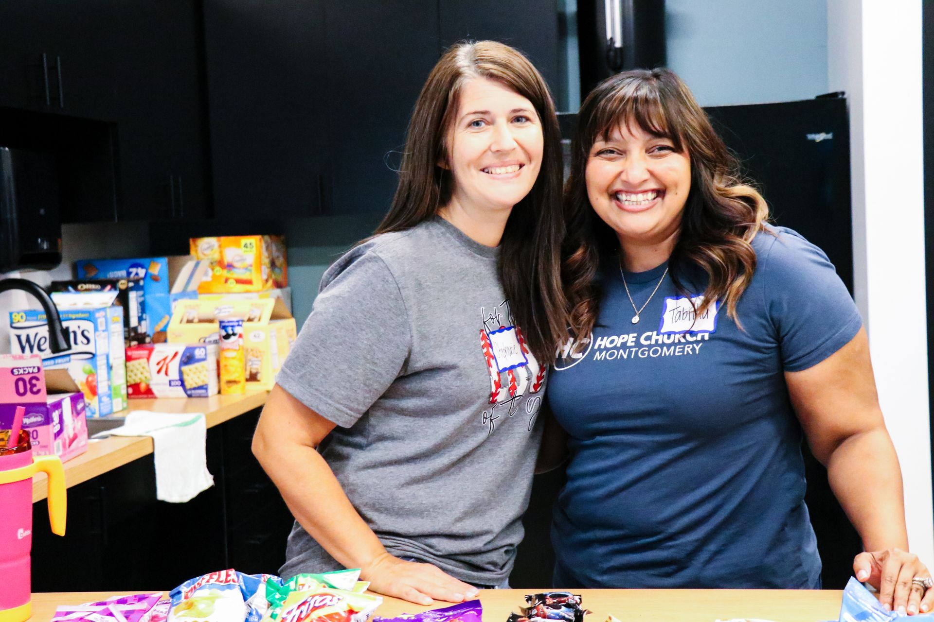 Two women smiling behind a table of colorful items at an indoor event