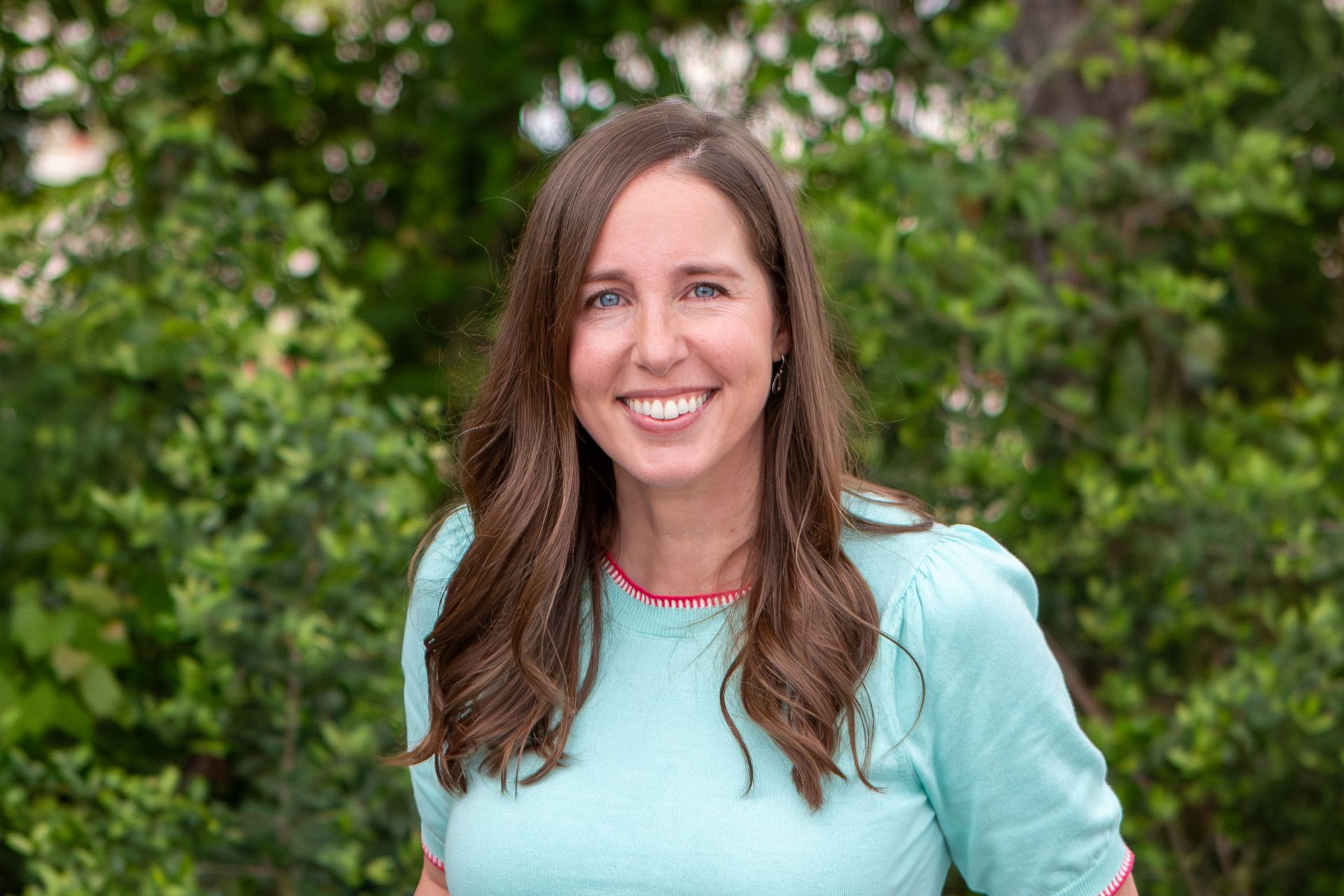 Smiling woman in a light blue top standing outdoors in front of green foliage
