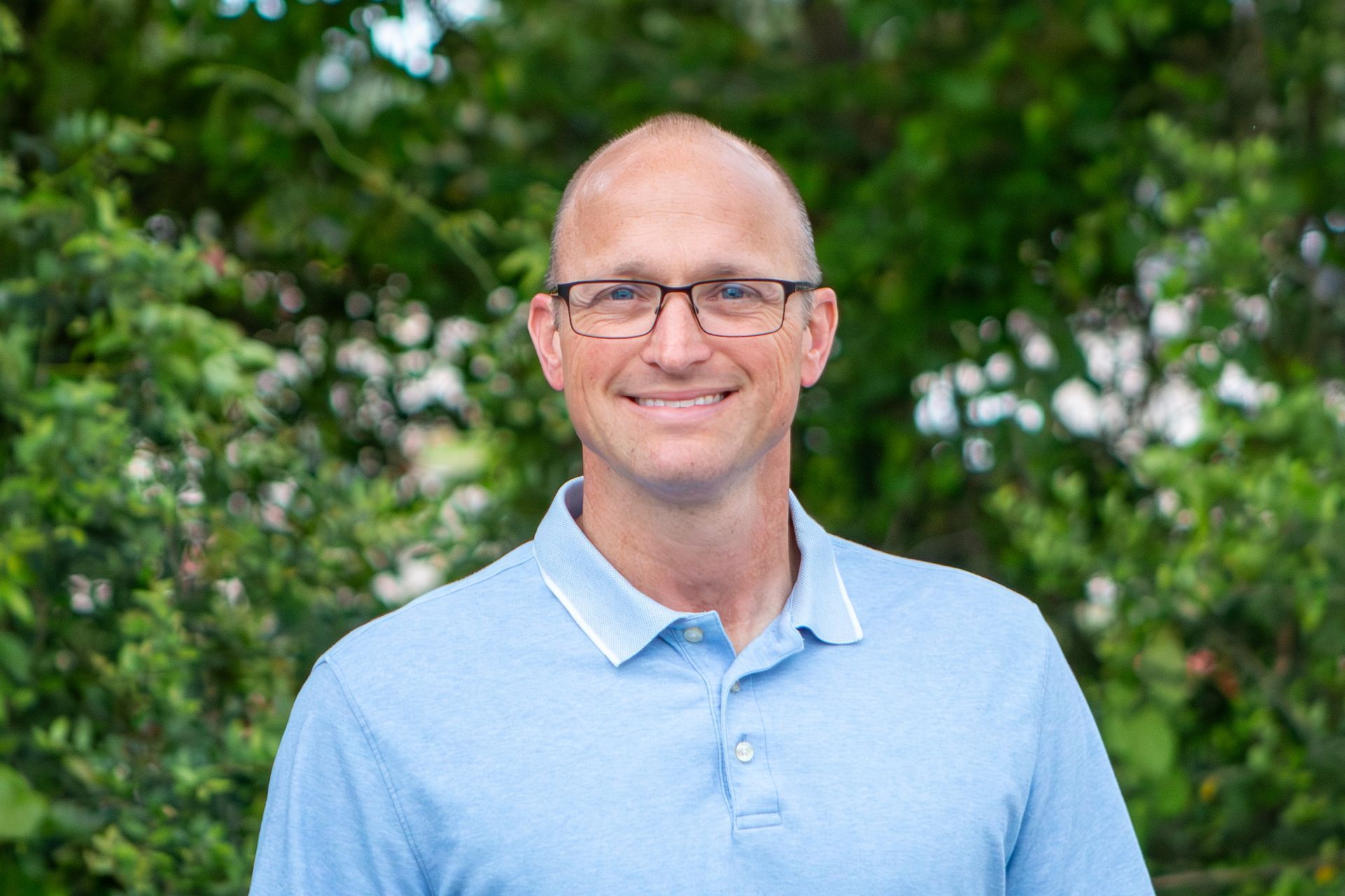 Smiling bald man in glasses wearing a light blue polo, standing outdoors with green foliage behind him