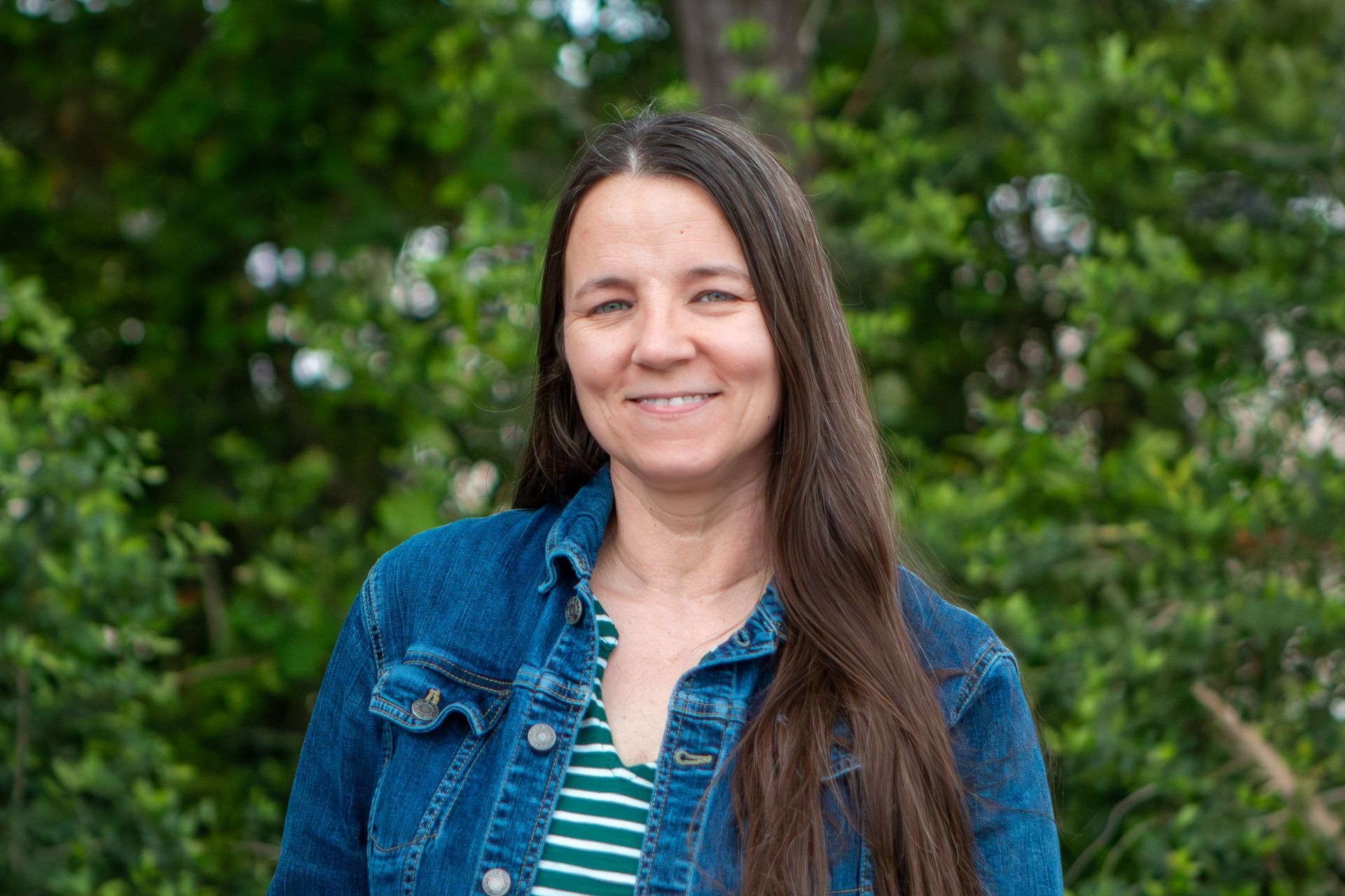 Woman in a denim jacket smiling outdoors with green trees in the background
