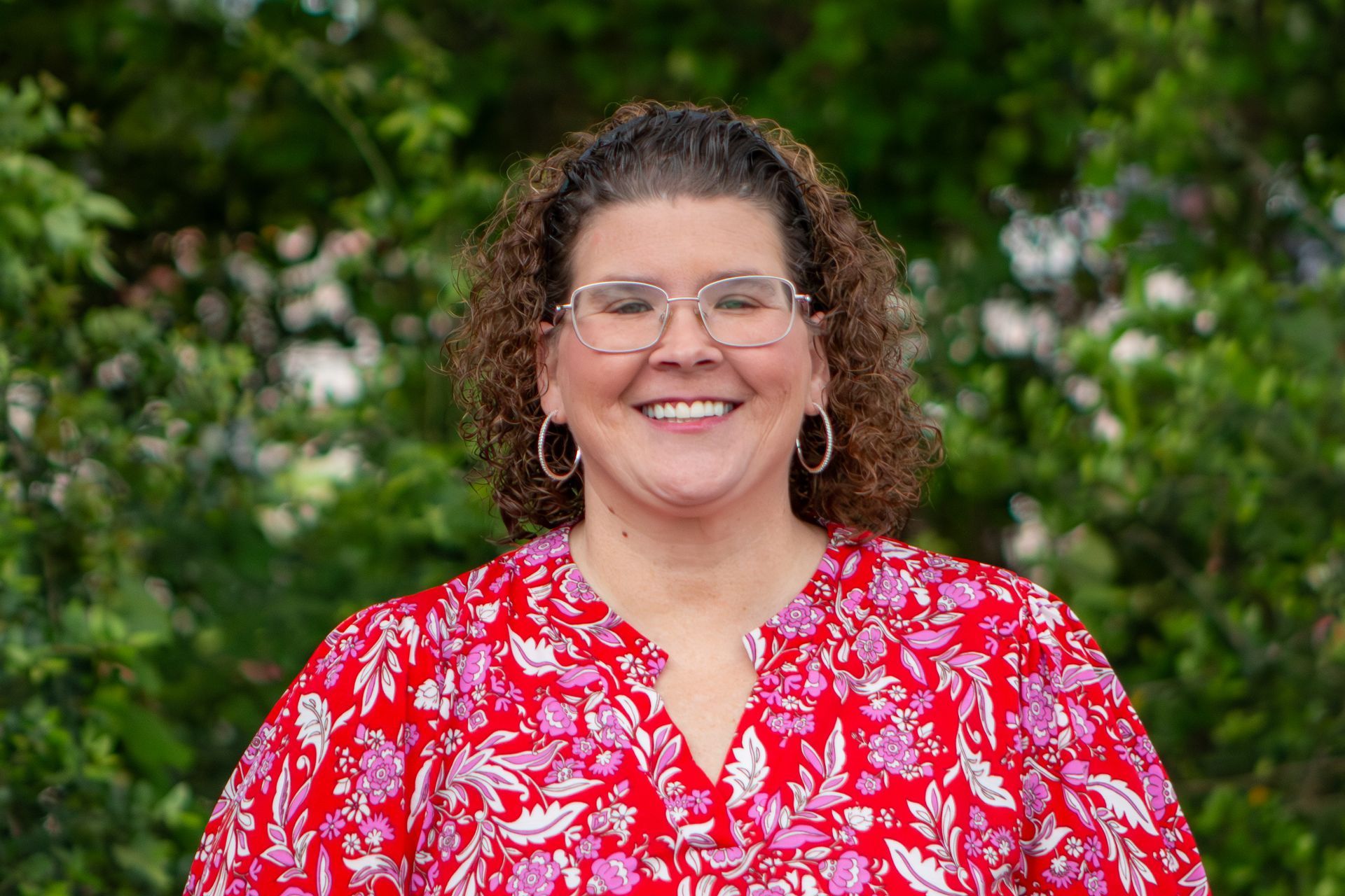 Smiling person in glasses and a red floral blouse standing outdoors with green foliage behind.
