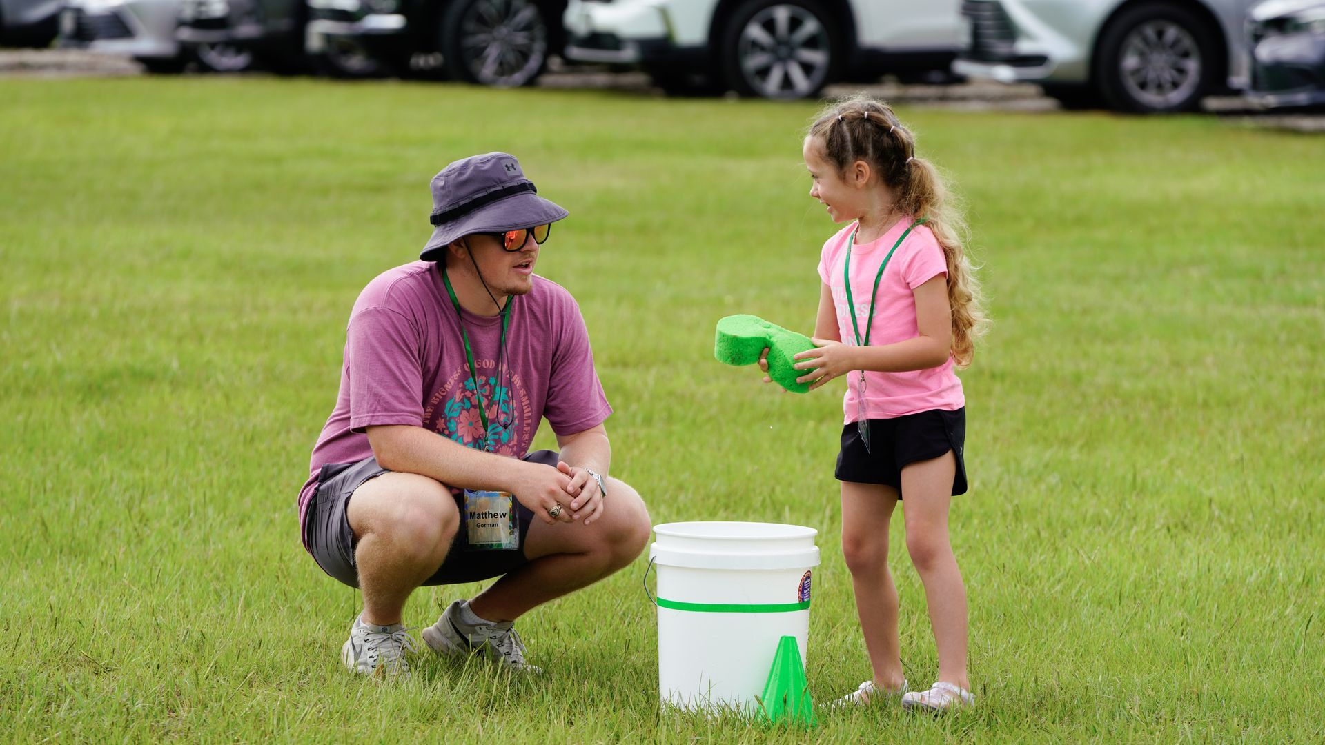 Adult crouches beside a child on a grassy field as they talk near a white bucket and green sponges.