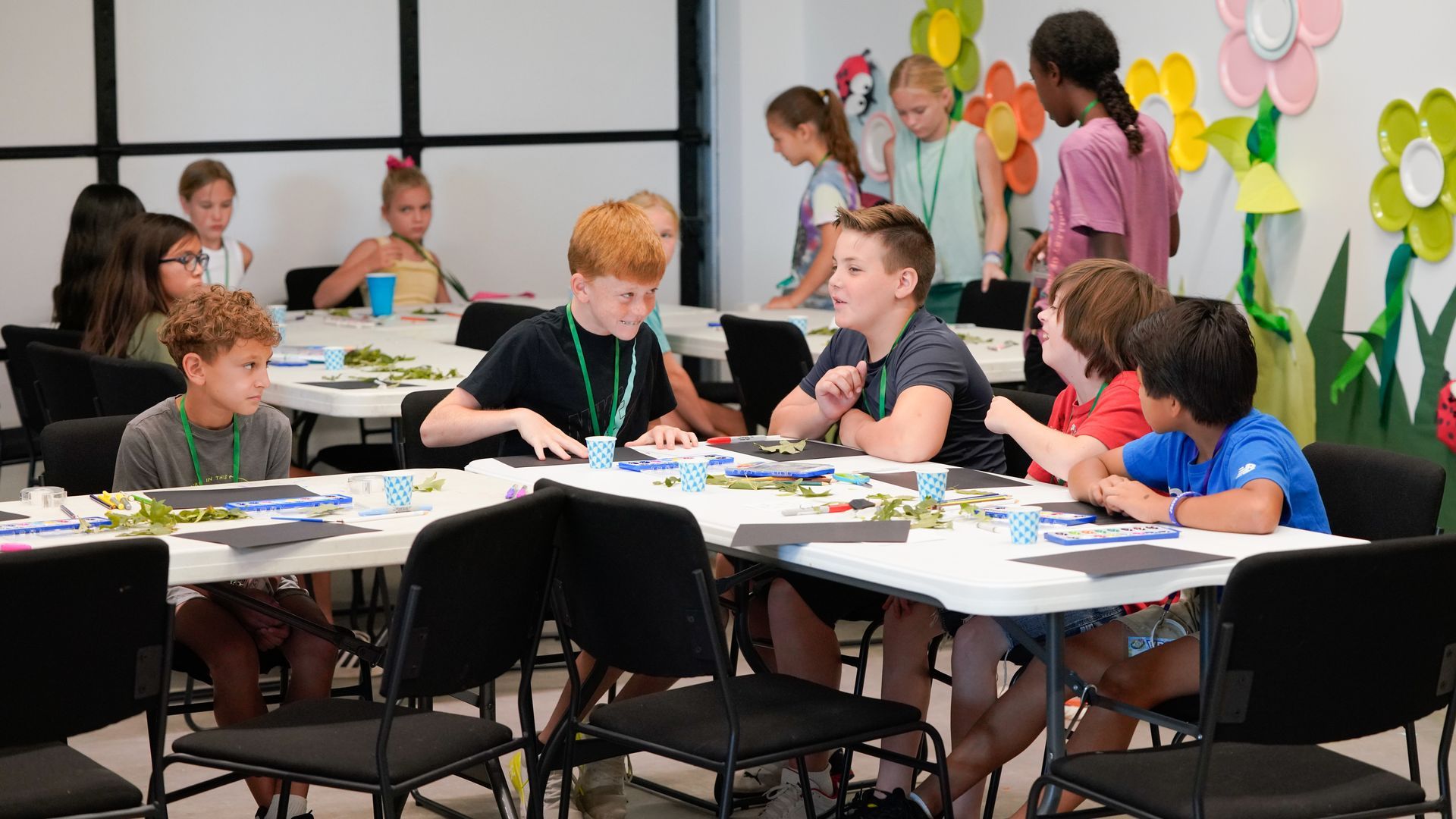 Students working at tables in a classroom, with colorful paper flowers on the wall.