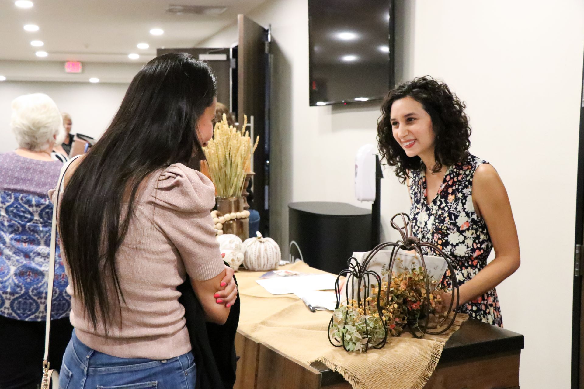 Two women chatting at a decorated market booth with pumpkins and autumn decor