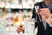 A person in a suit prepares a cocktail, holding a jigger over a glass of ice at a bar.