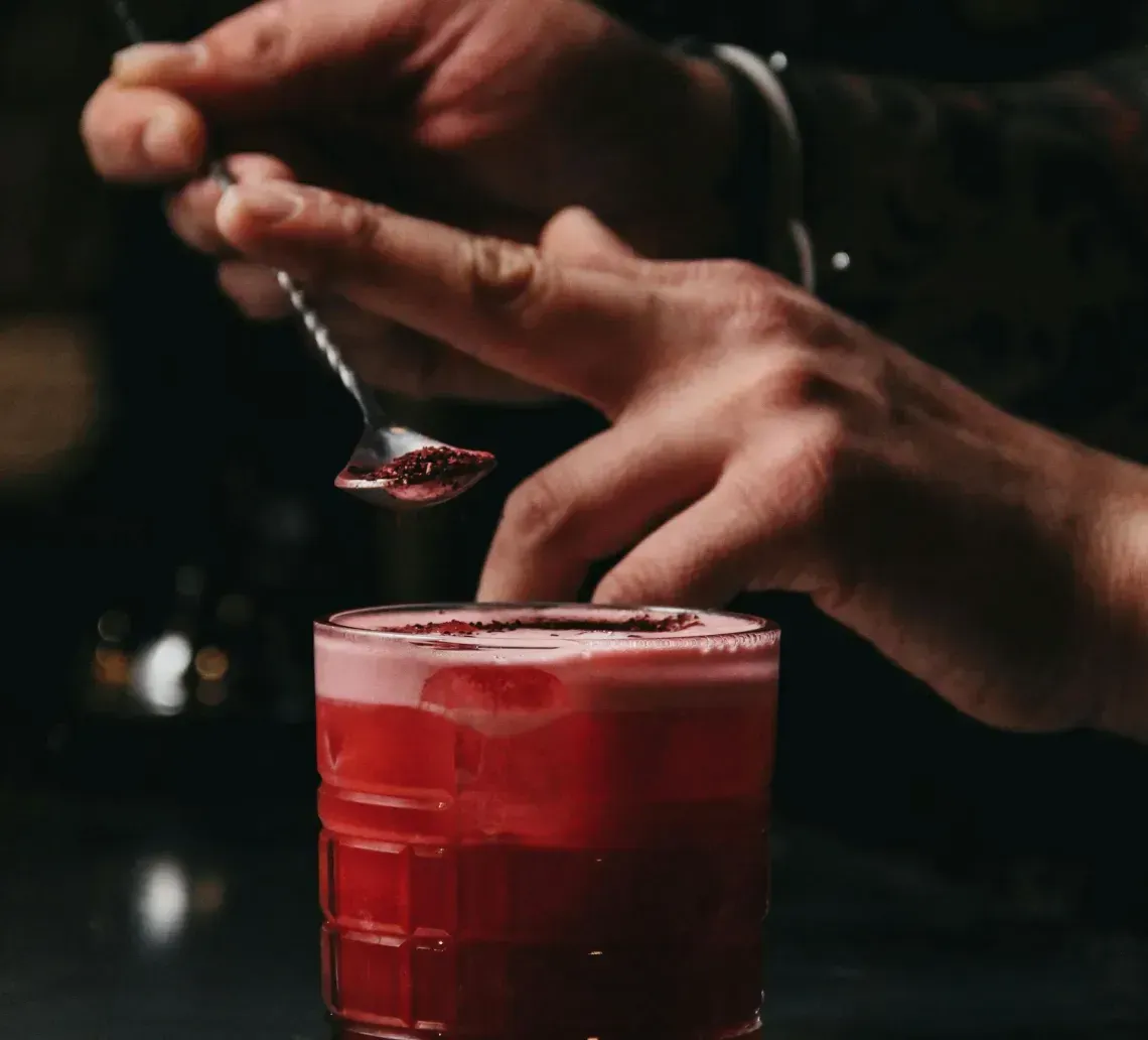 A bartender holds a long spoon over a vibrant red cocktail in a textured glass on a dimly lit bar counter.