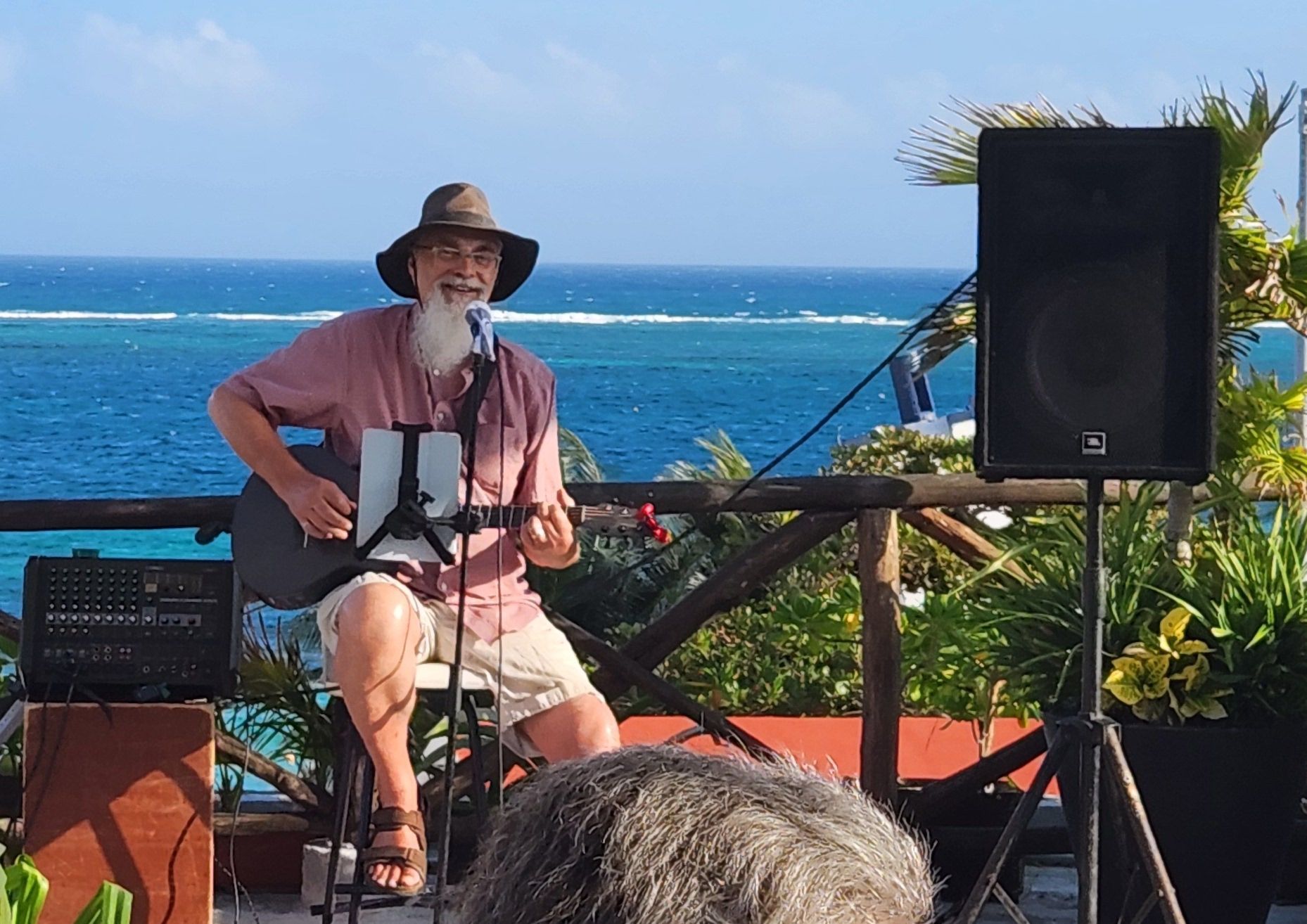 Randy smiling and playing acoustic guitar on the rooftop, where beautiful blue waters are waving right behind him.