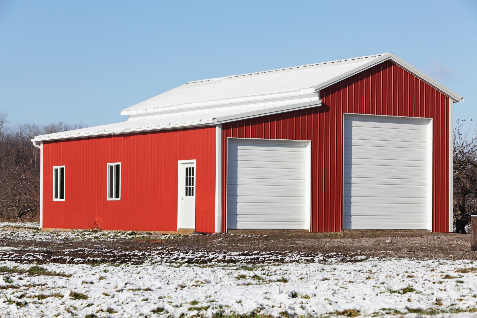 A red barn with two white garage doors is in the middle of a snowy field.