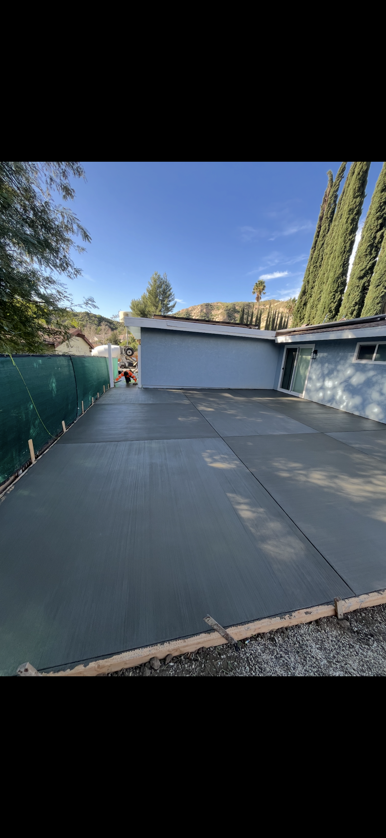 Freshly poured concrete driveway in front of a blue house and green fence.