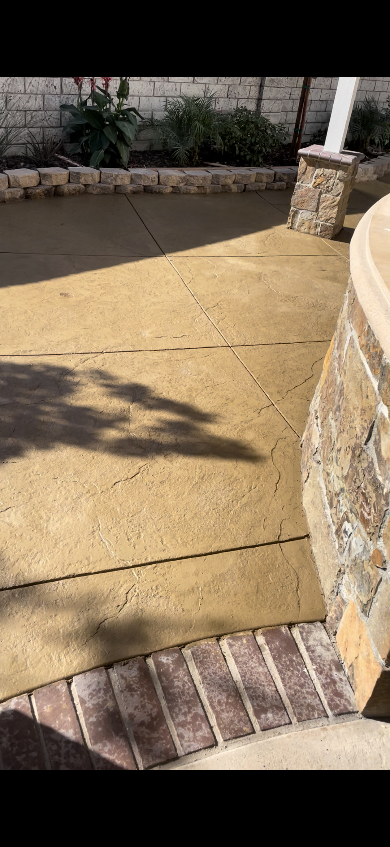 Sunlit patio with stone retaining wall and brick edging. Shadow of tree casts across the surface.