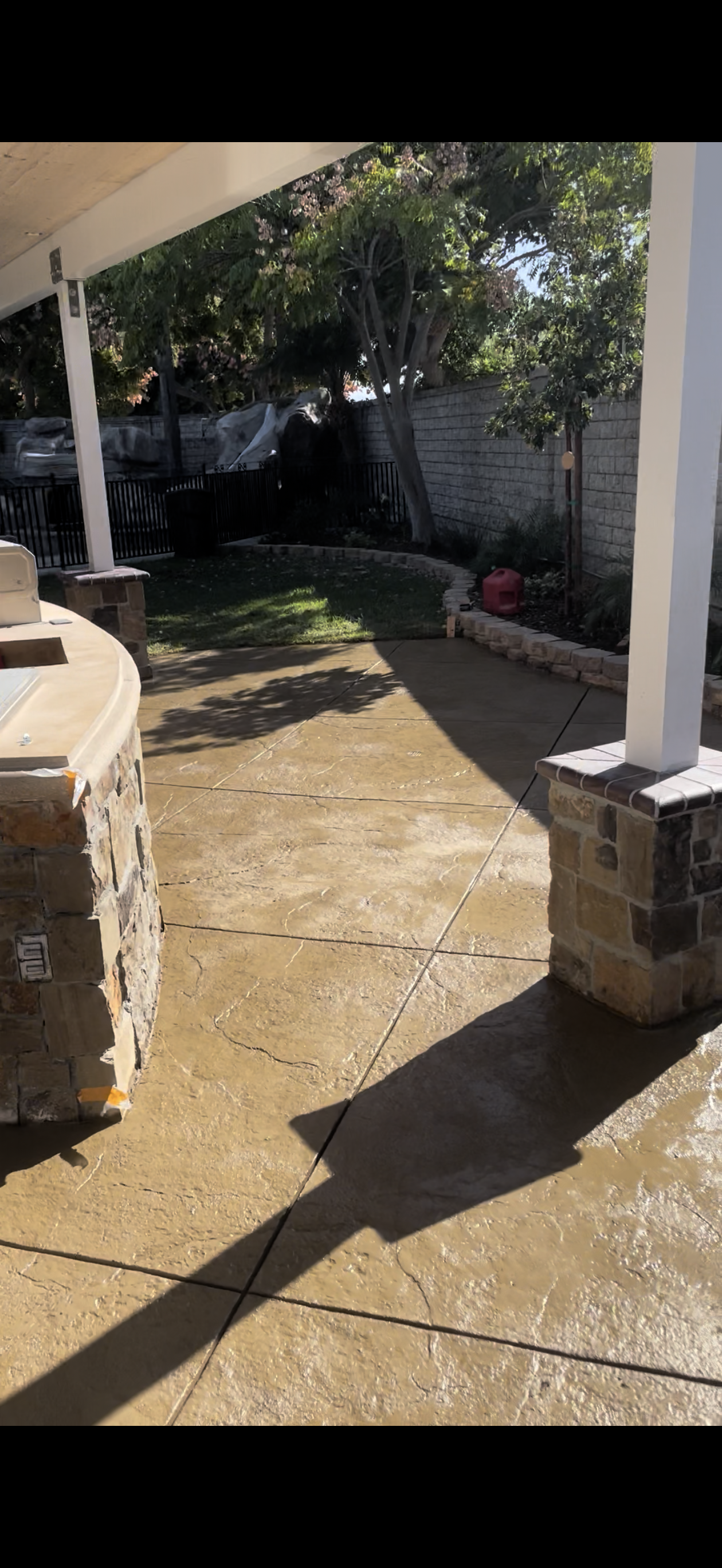 Patio with a stone structure, concrete pathway, and backyard greenery. Bright sunlight creates long shadows.