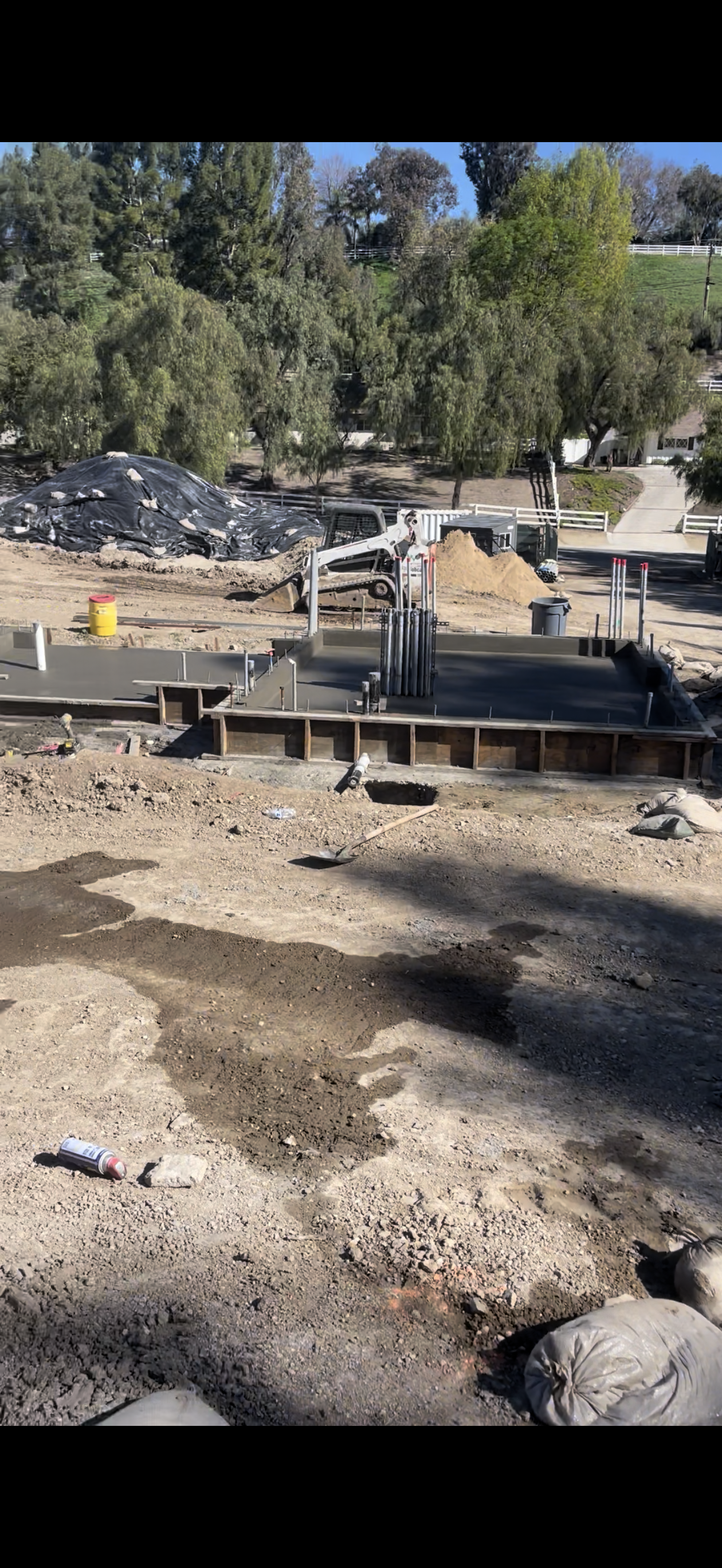 Construction site with concrete forms, a pile of material, and trees in the background.