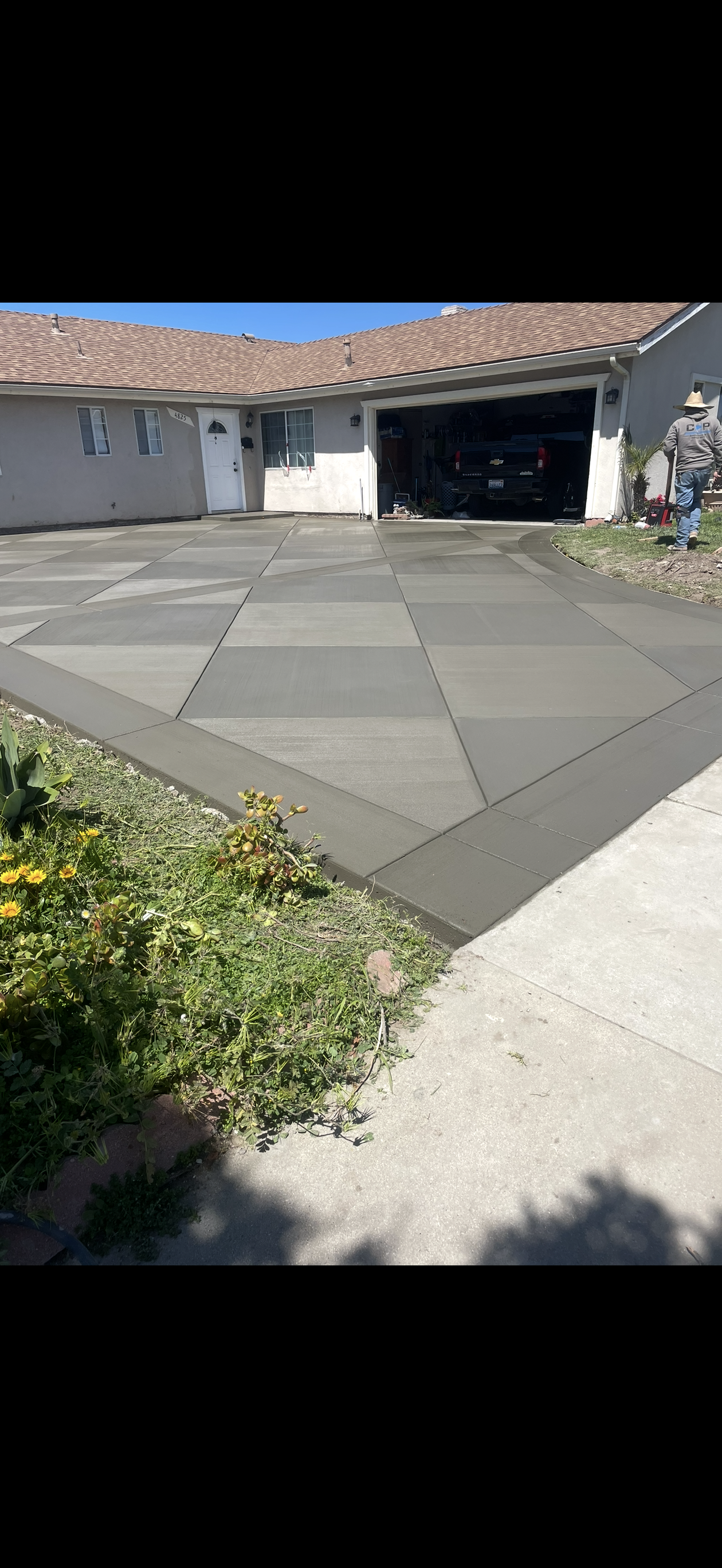 A newly paved concrete driveway in front of a house. Bordered by green bushes and a sidewalk.