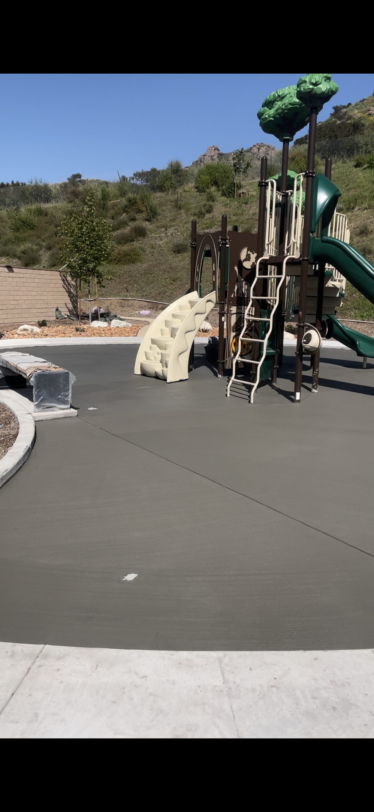 Playground with green slides, a climbing structure, and gray surface. Hills and blue sky in the background.