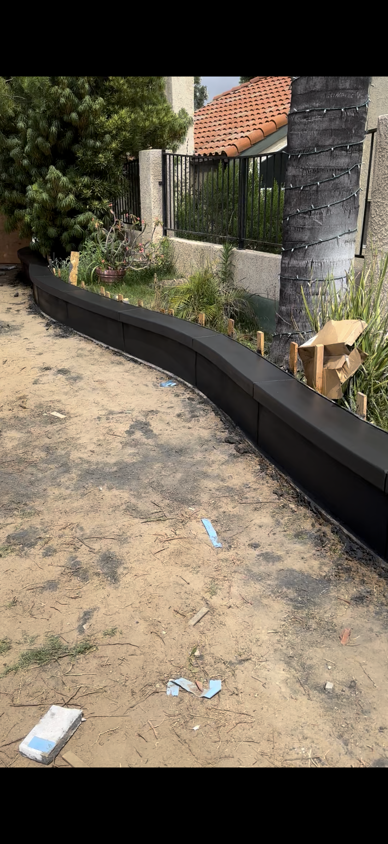 A curved black border along a sandy area with weeds, a building in the background.