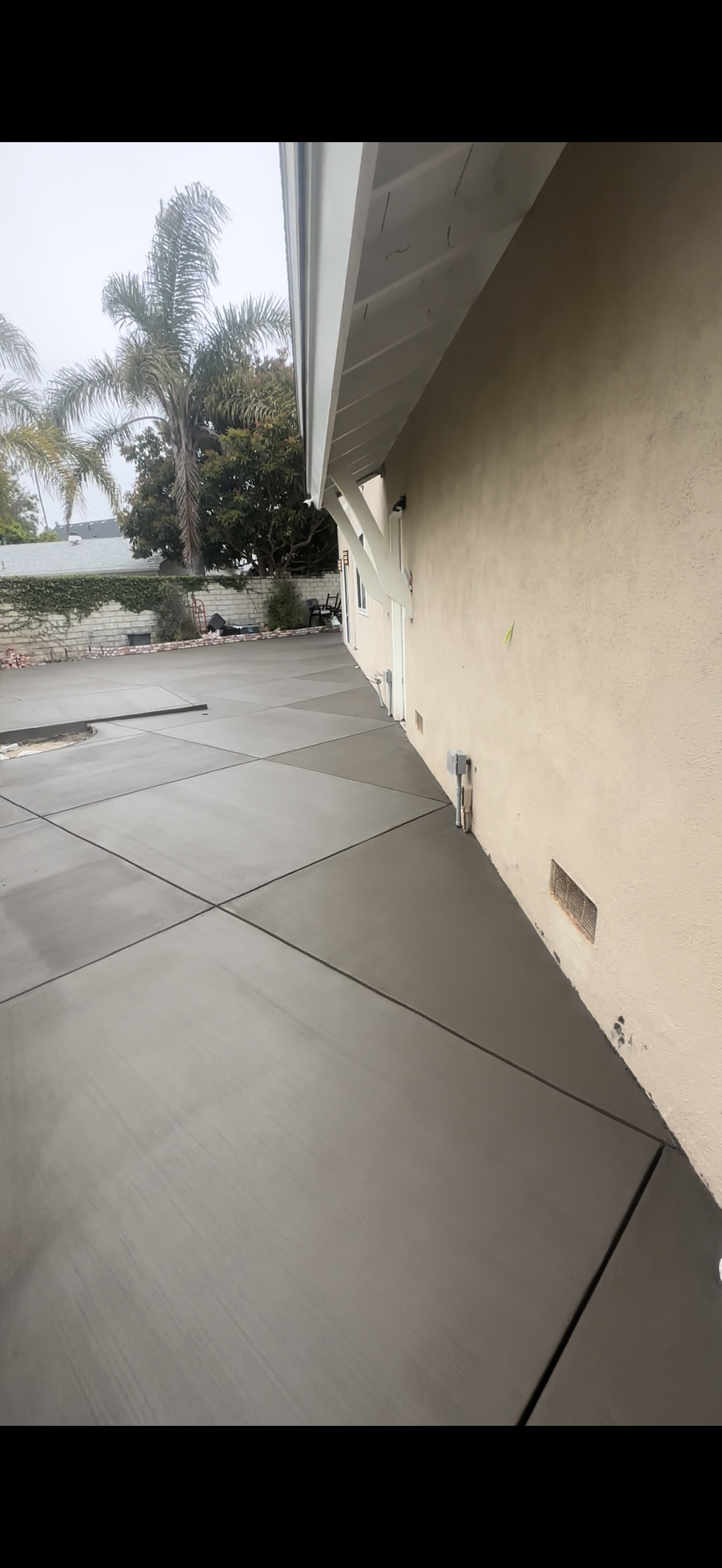Newly poured concrete patio next to a light-colored stucco building, cloudy sky in the background.