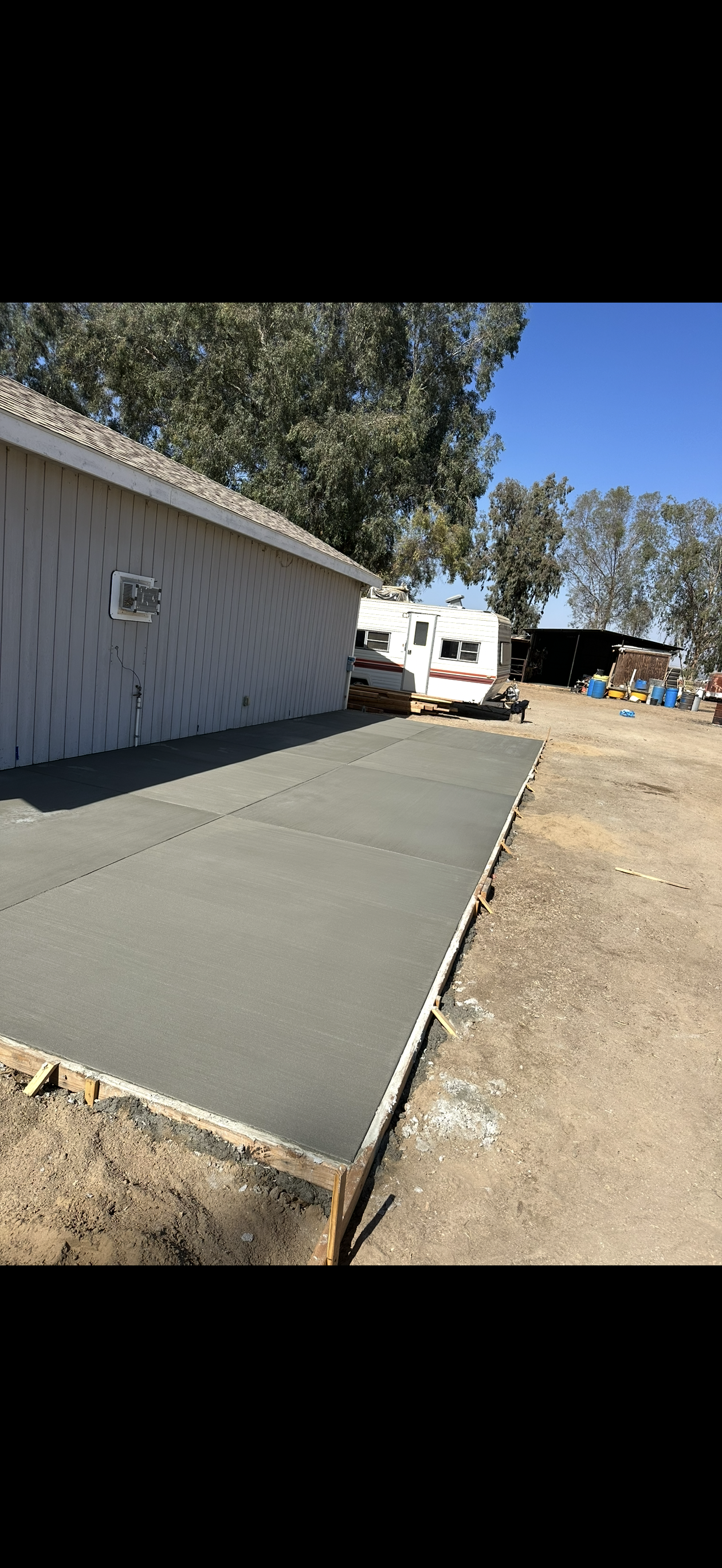 Freshly poured concrete patio next to a light-colored building, with a trailer and trees in the background.