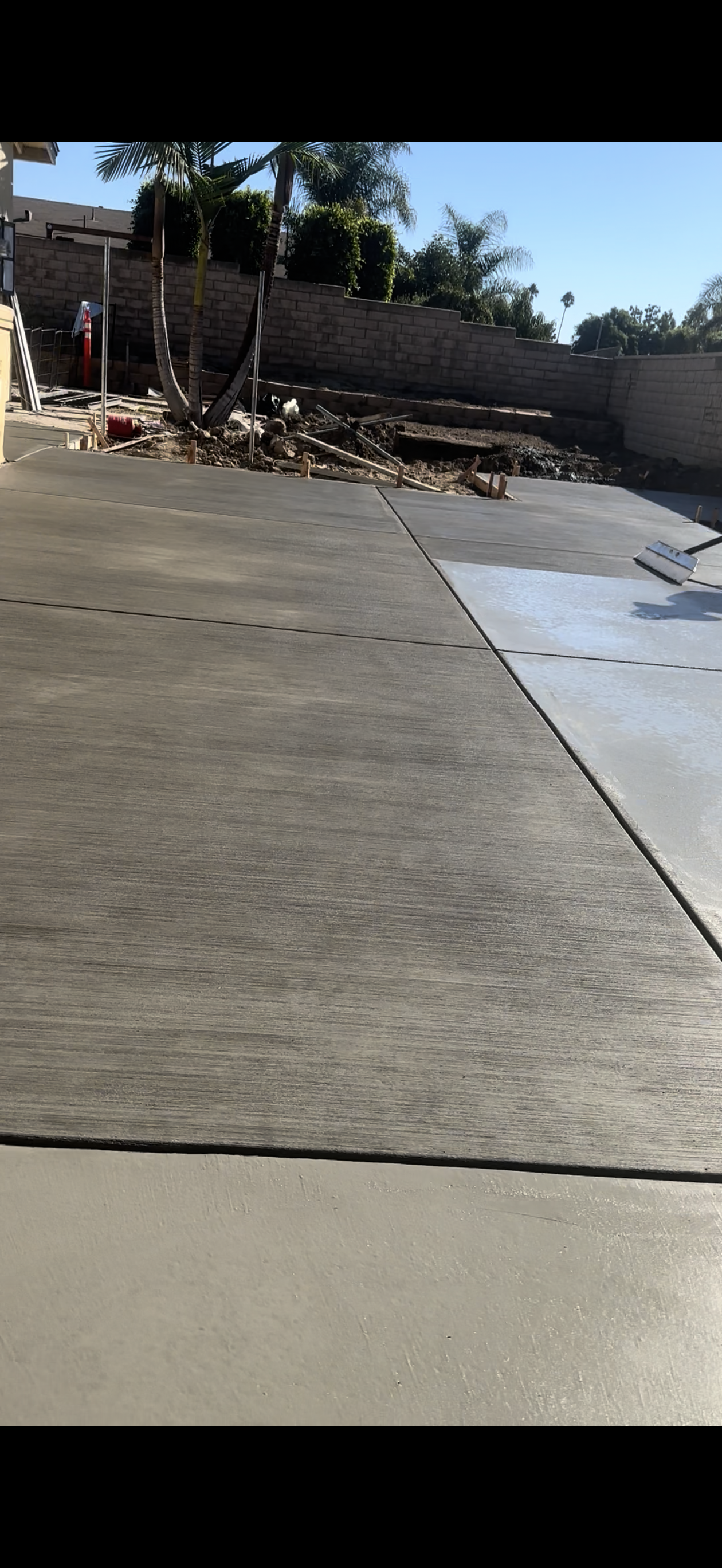 Newly poured concrete patio, part of which is drying, with a debris-filled area and a retaining wall in the background.
