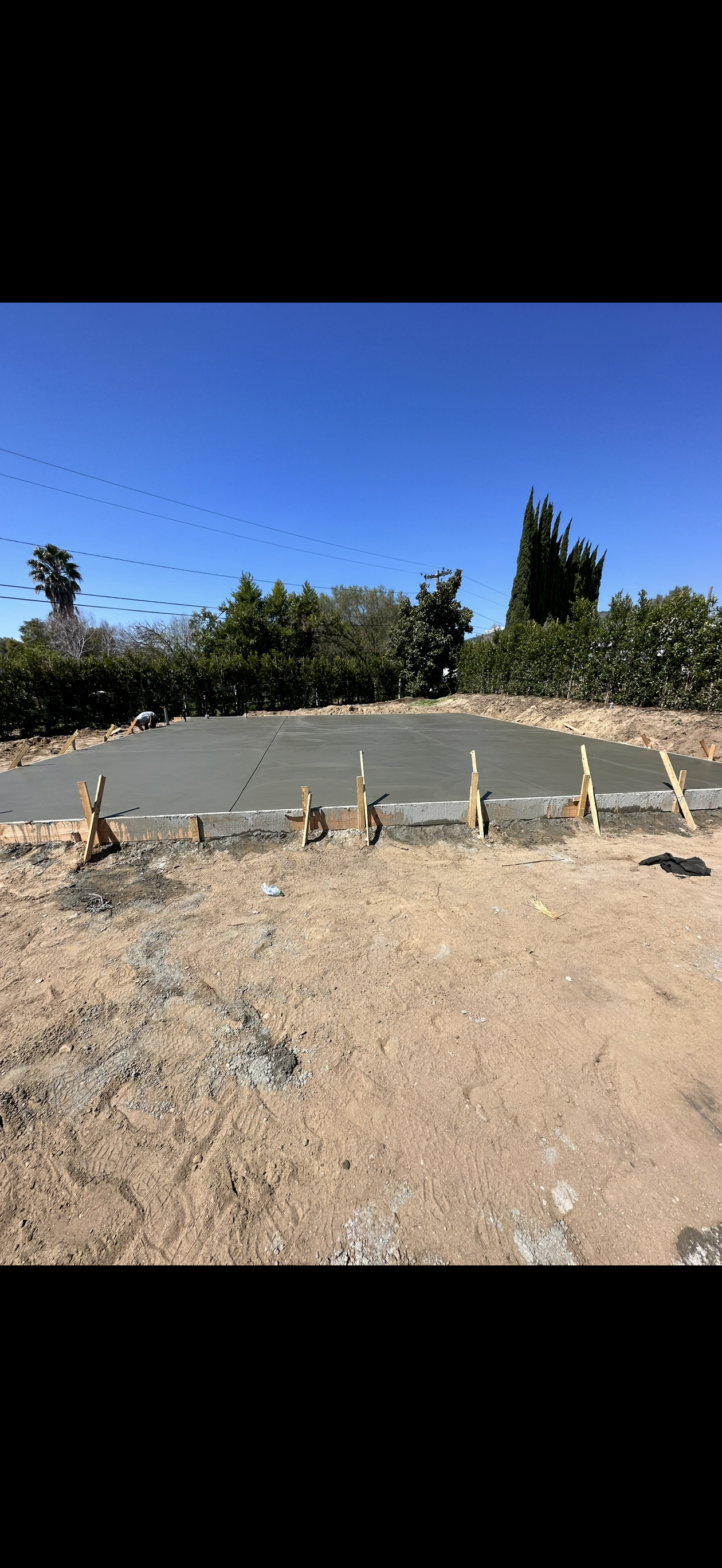 Freshly poured concrete slab with wooden supports on a dirt lot under a blue sky.