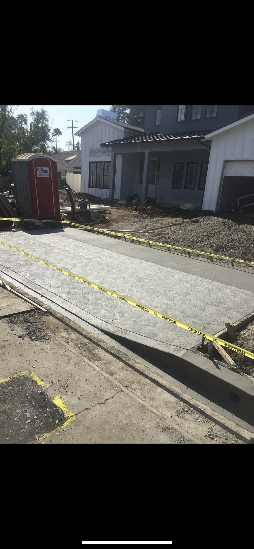 Road construction site with house in the background. Yellow caution tape and a portable toilet are visible.