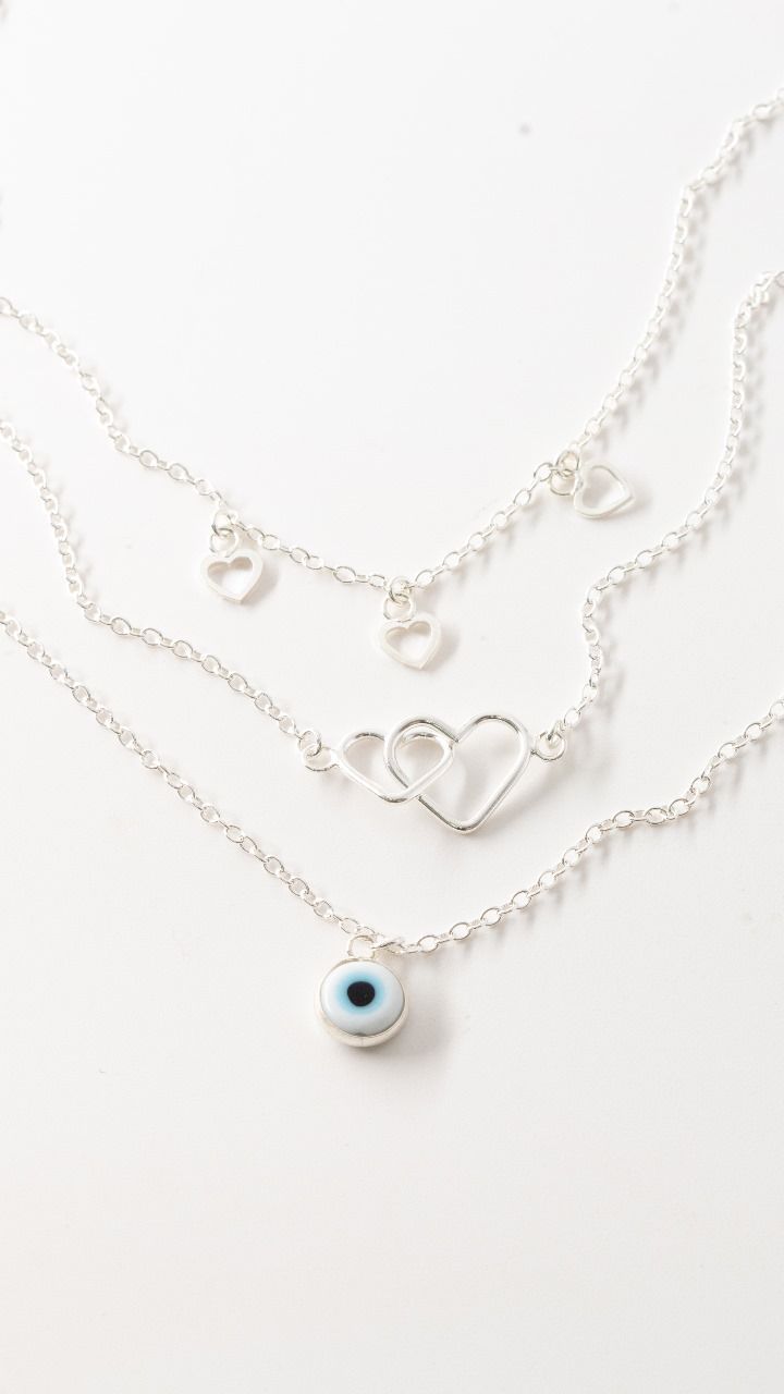 Tres collares de plata superpuestos con dijes de corazón y un colgante de ojo azul sobre un fondo blanco.
