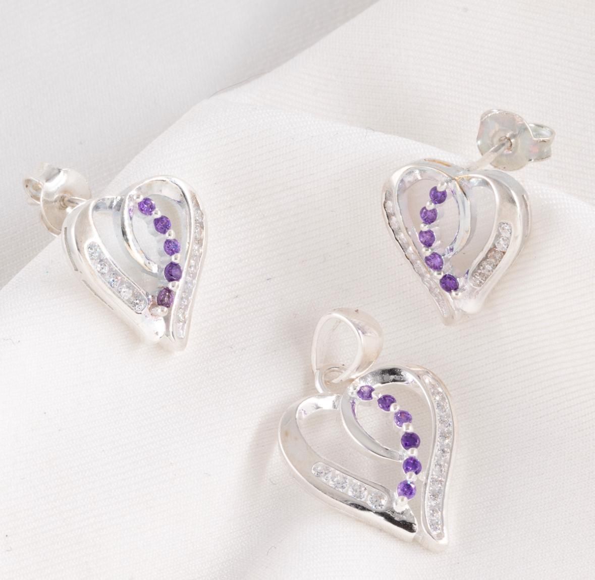 Tres colgantes plateados en forma de corazón con piedras moradas y transparentes sobre un fondo de tela blanca.