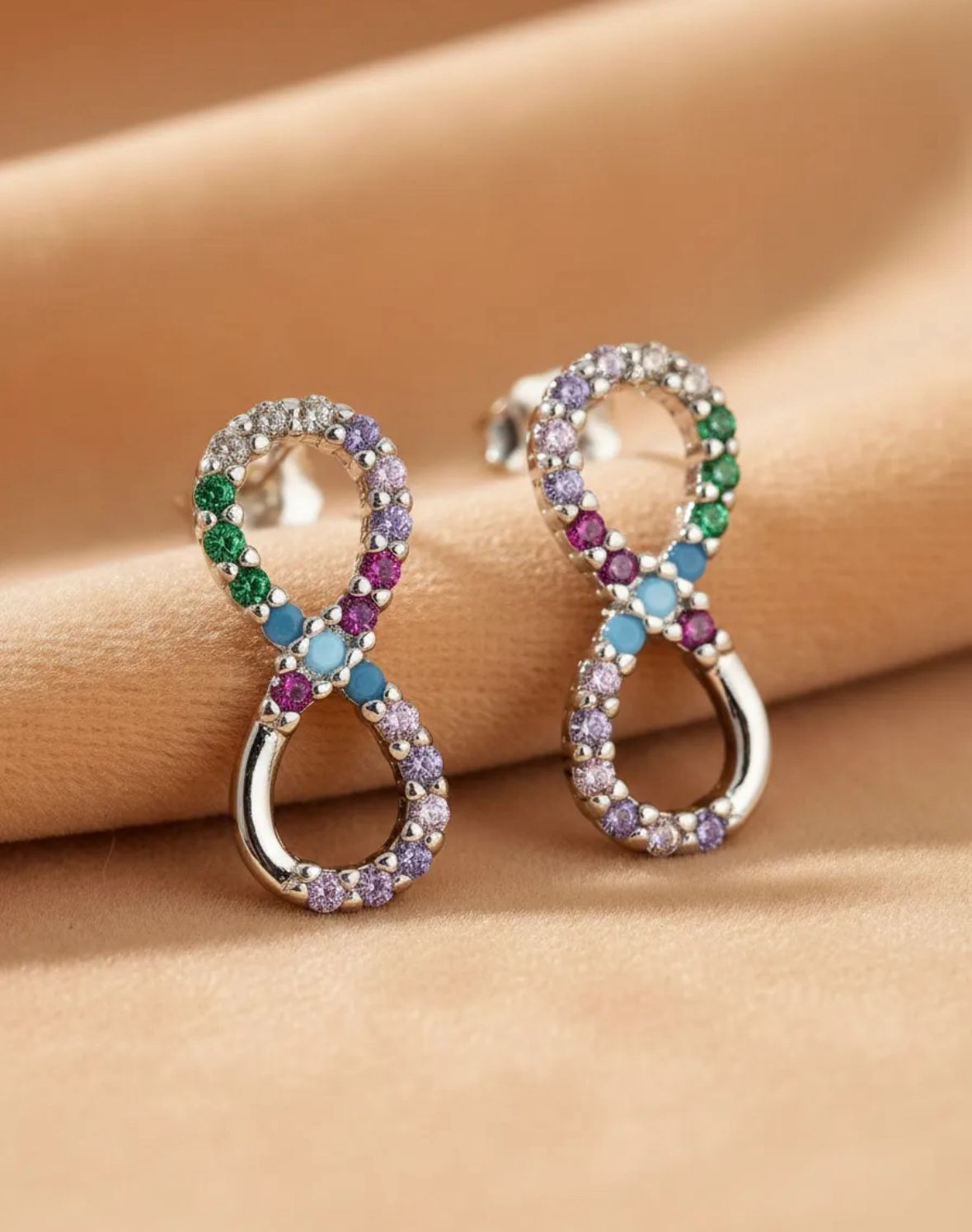 Dos pendientes de infinito con piedras preciosas verdes, moradas y transparentes sobre un fondo color canela.