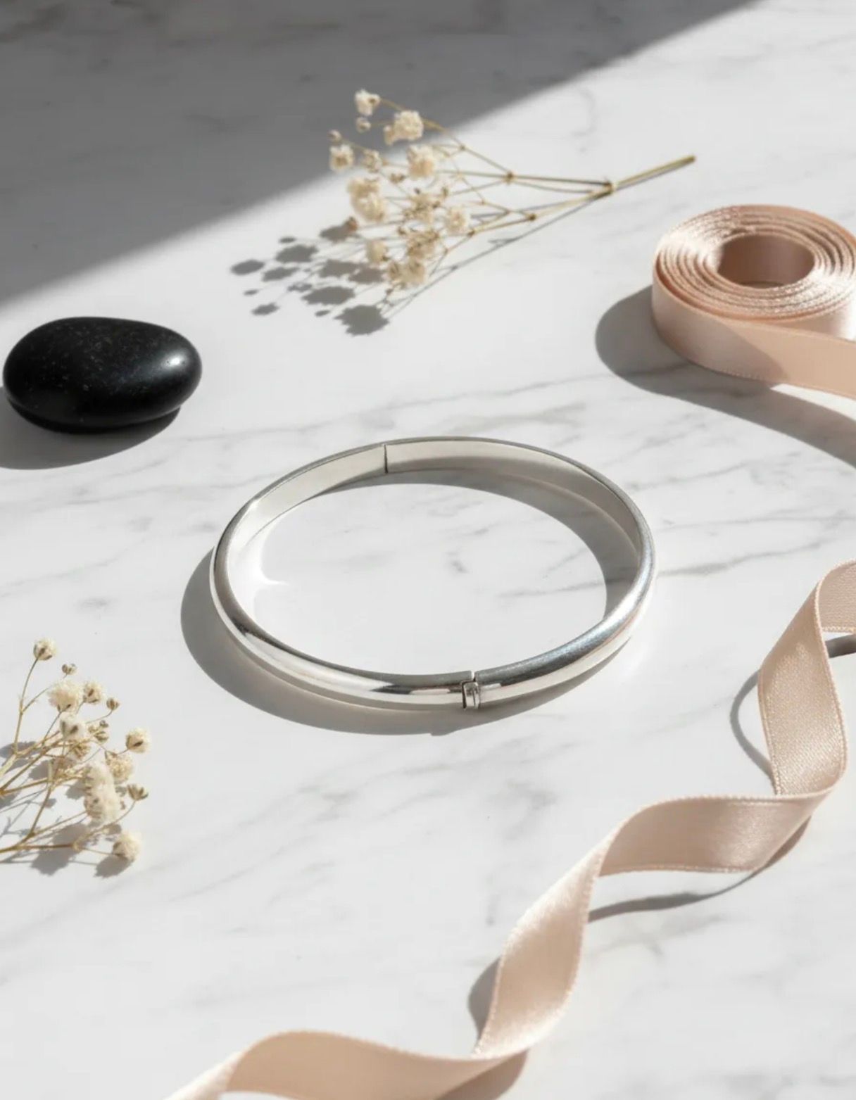 Anillo redondo de metal y cinta beige sobre una superficie de mármol blanco con una piedra negra y flores secas.
