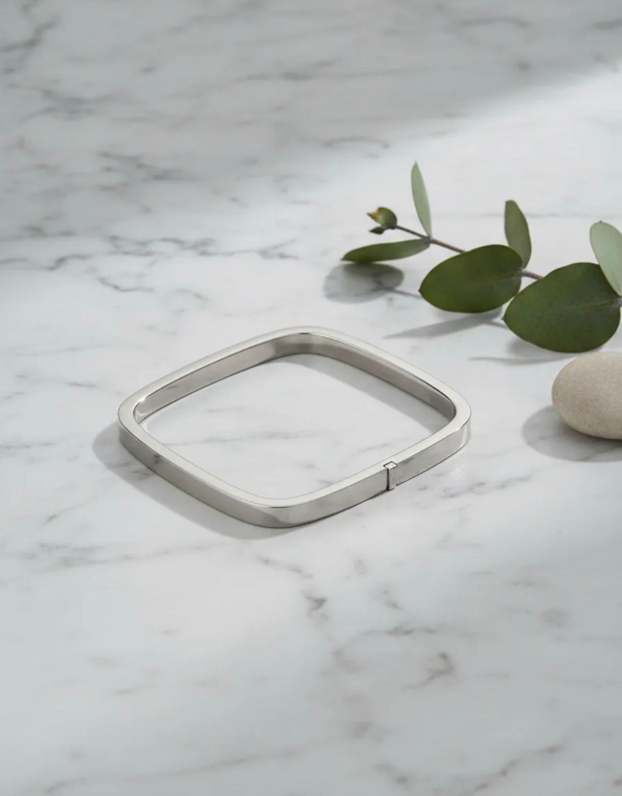 Pulsera cuadrada de plata sobre una superficie de mármol blanco con hojas de eucalipto cerca.