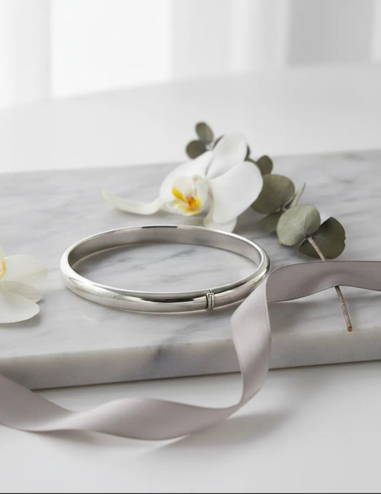 Brazalete de plata sobre una caja blanca con cinta de satén y flores de orquídea blanca.