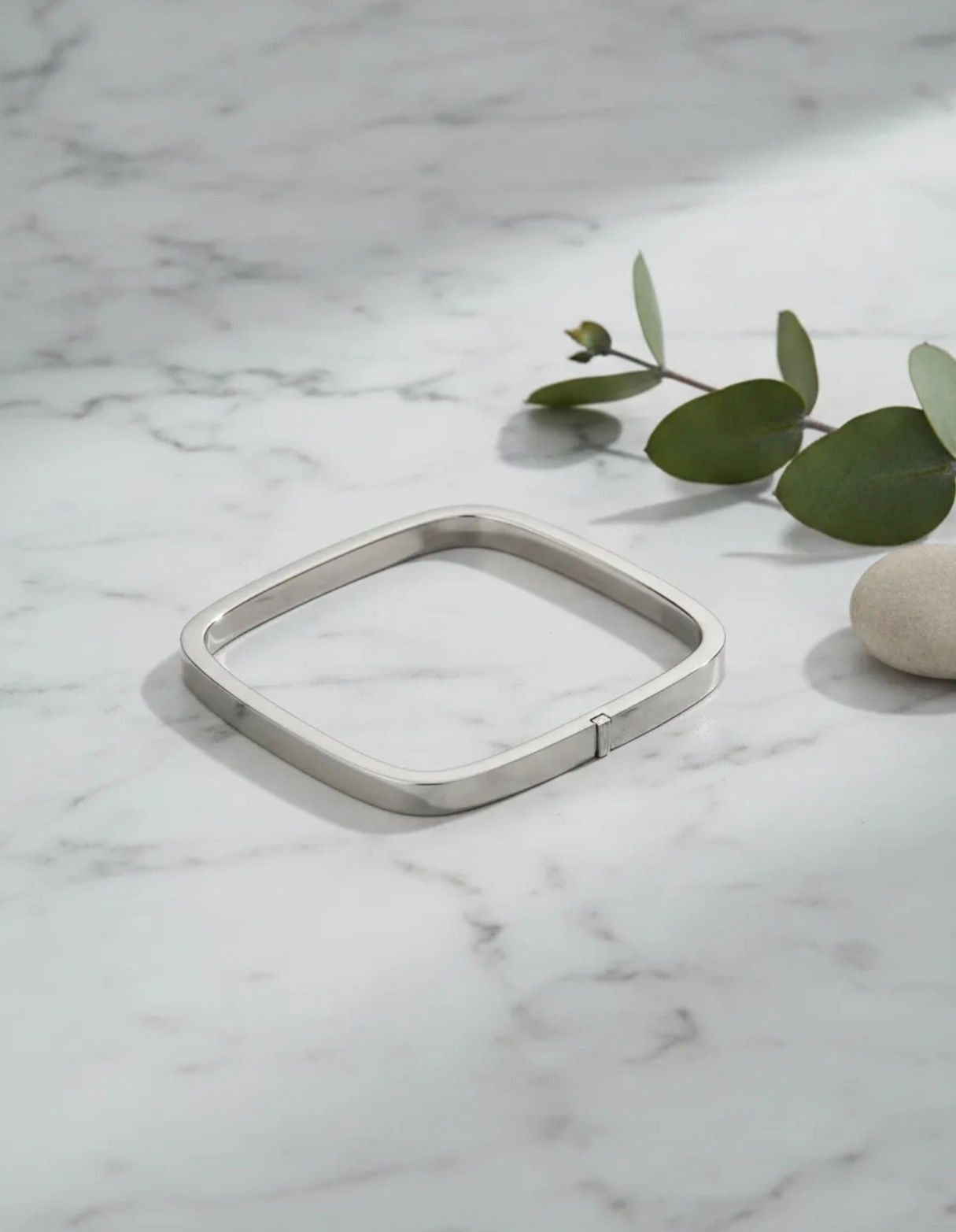 Pulsera cuadrada de plata sobre mármol blanco junto a hojas verdes y una pequeña piedra.
