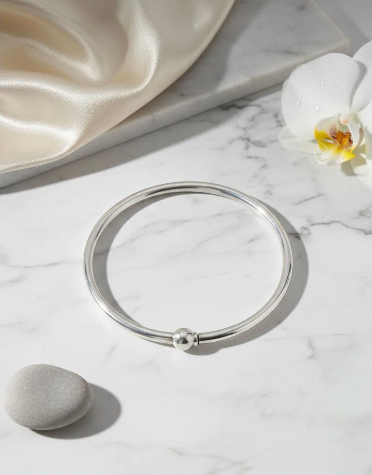 Pulsera de plata sobre una superficie de mármol con tela de seda, una piedra blanca y una flor de orquídea.