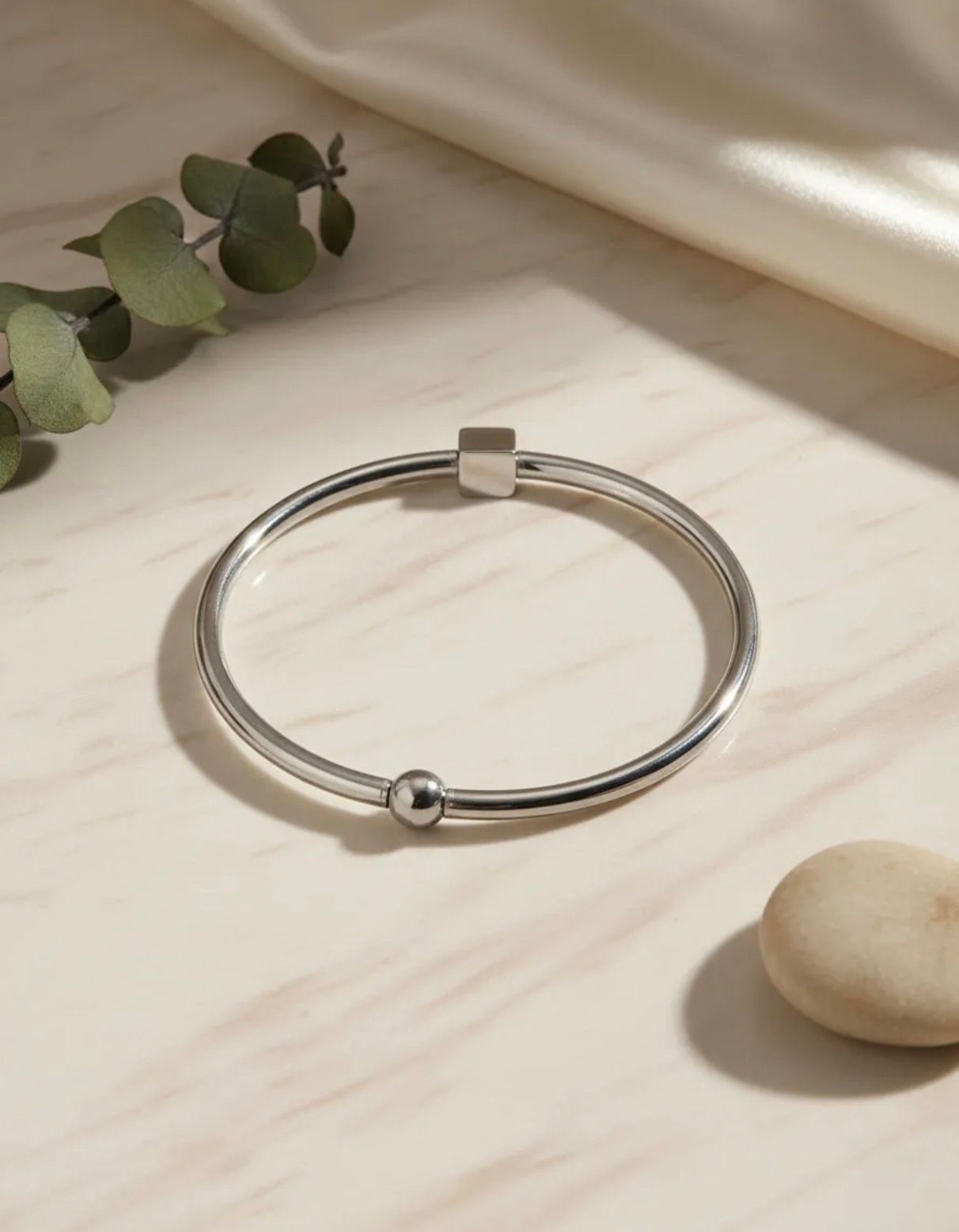 Pulsera de plata con una pequeña cuenta, sobre una superficie de mármol con hojas de eucalipto cerca.