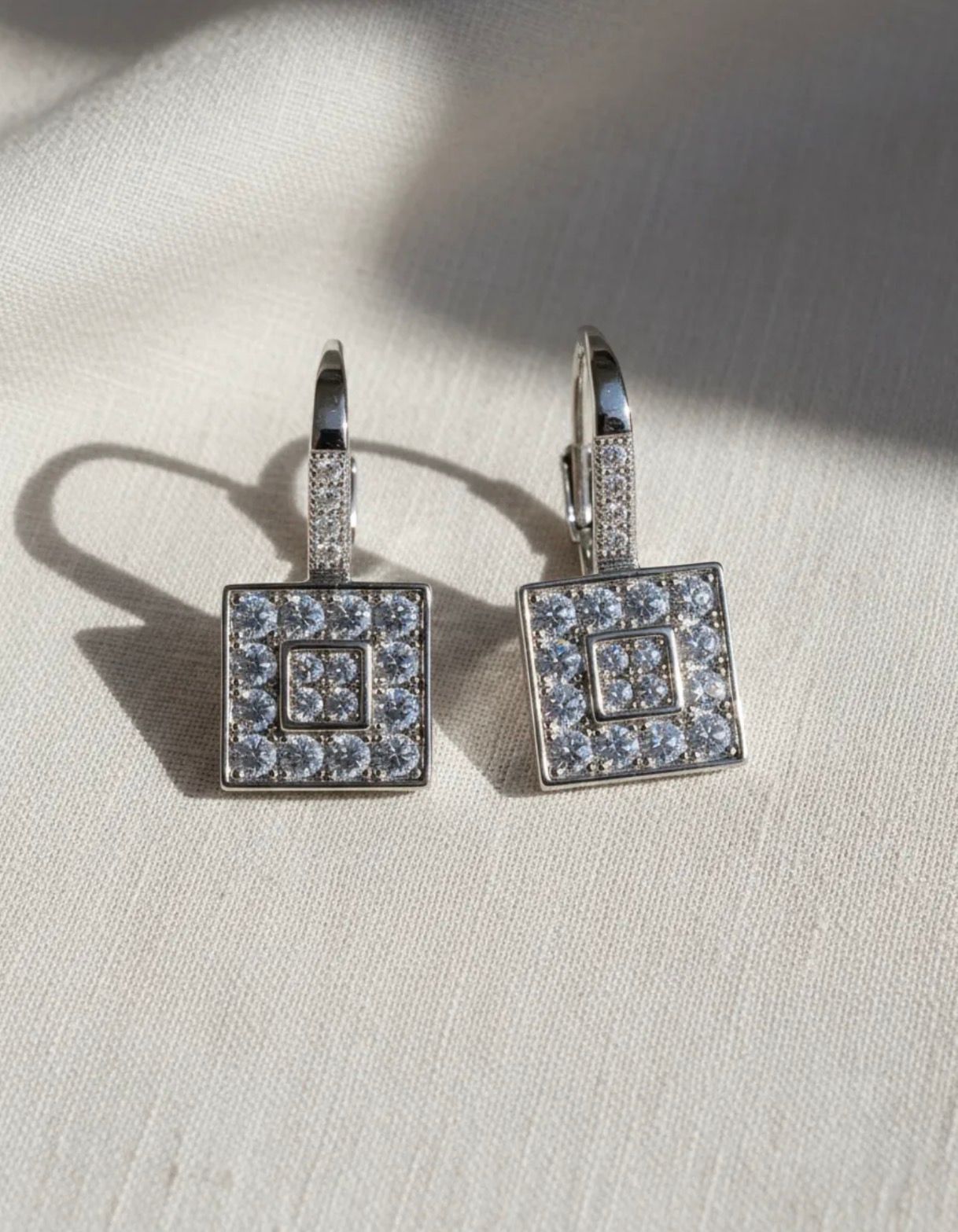 Par de pendientes colgantes cuadrados con incrustaciones de diamantes sobre un fondo de tela clara.
