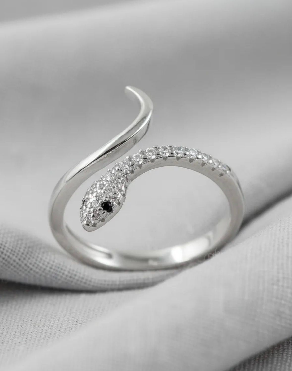 Anillo plateado con forma de serpiente y cabeza curva incrustada de cristales sobre fondo de tela gris.