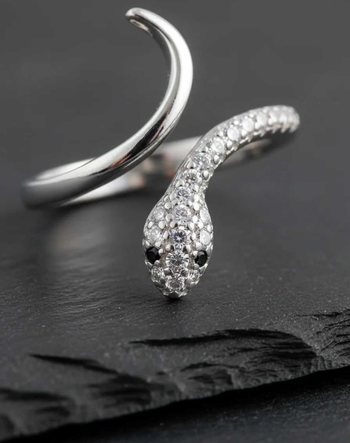 Anillo plateado con forma de serpiente y cabeza curva incrustada de cristales sobre fondo de tela gris.