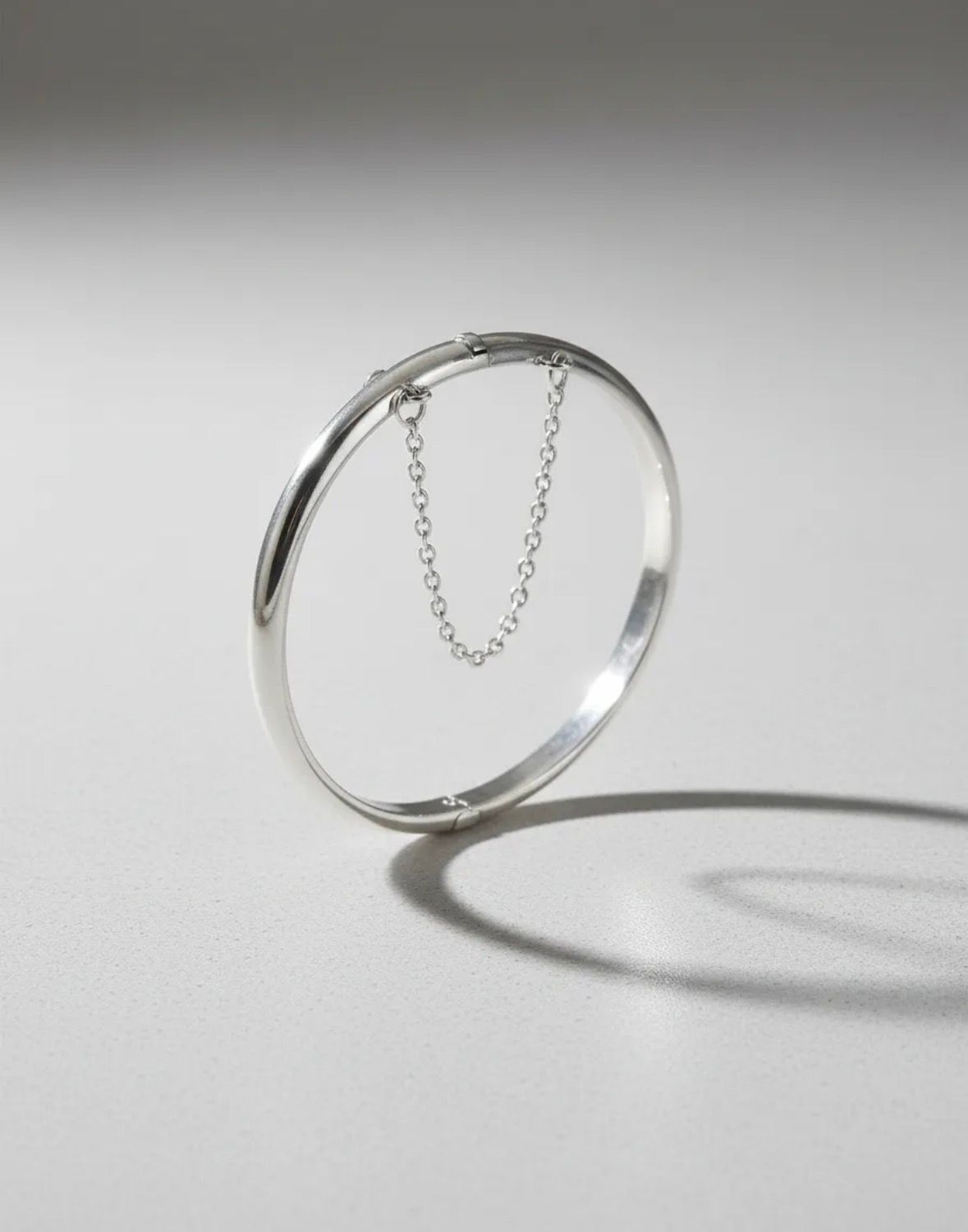 Anillo abierto de plata con una sombra de cadena colgante sobre un fondo claro.