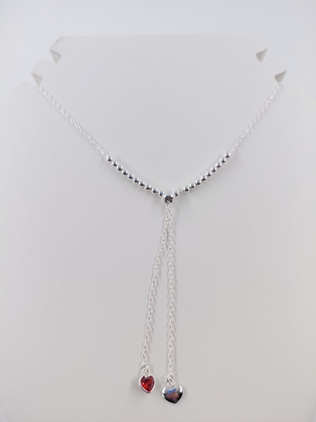 Collar de plata con colgantes en forma de corazón, uno rojo y otro iridiscente, sobre un fondo blanco.