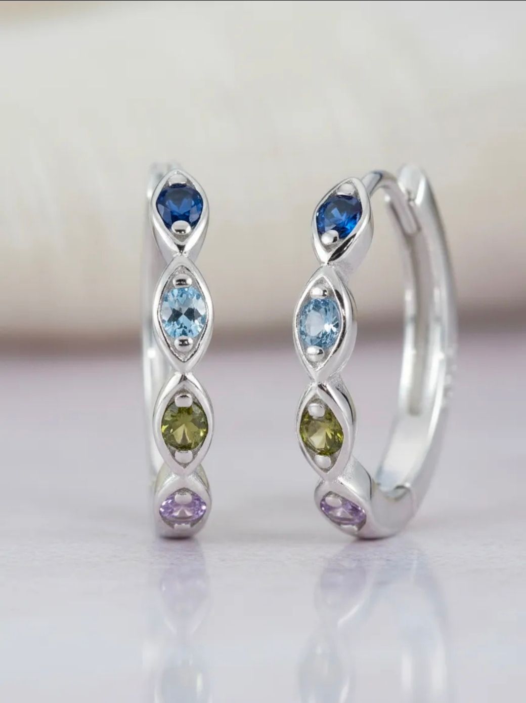 Dos pendientes de aro de plata con piedras preciosas azules, verdes y moradas sobre un fondo claro.