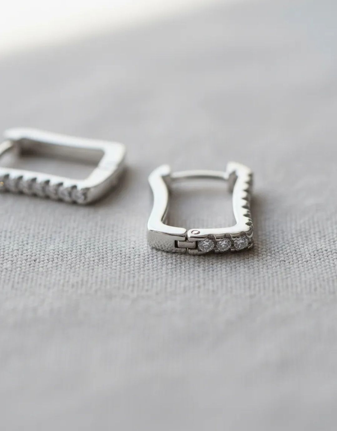 Dos pendientes rectangulares de plata con pequeños diamantes de imitación sobre una superficie de tela gris.